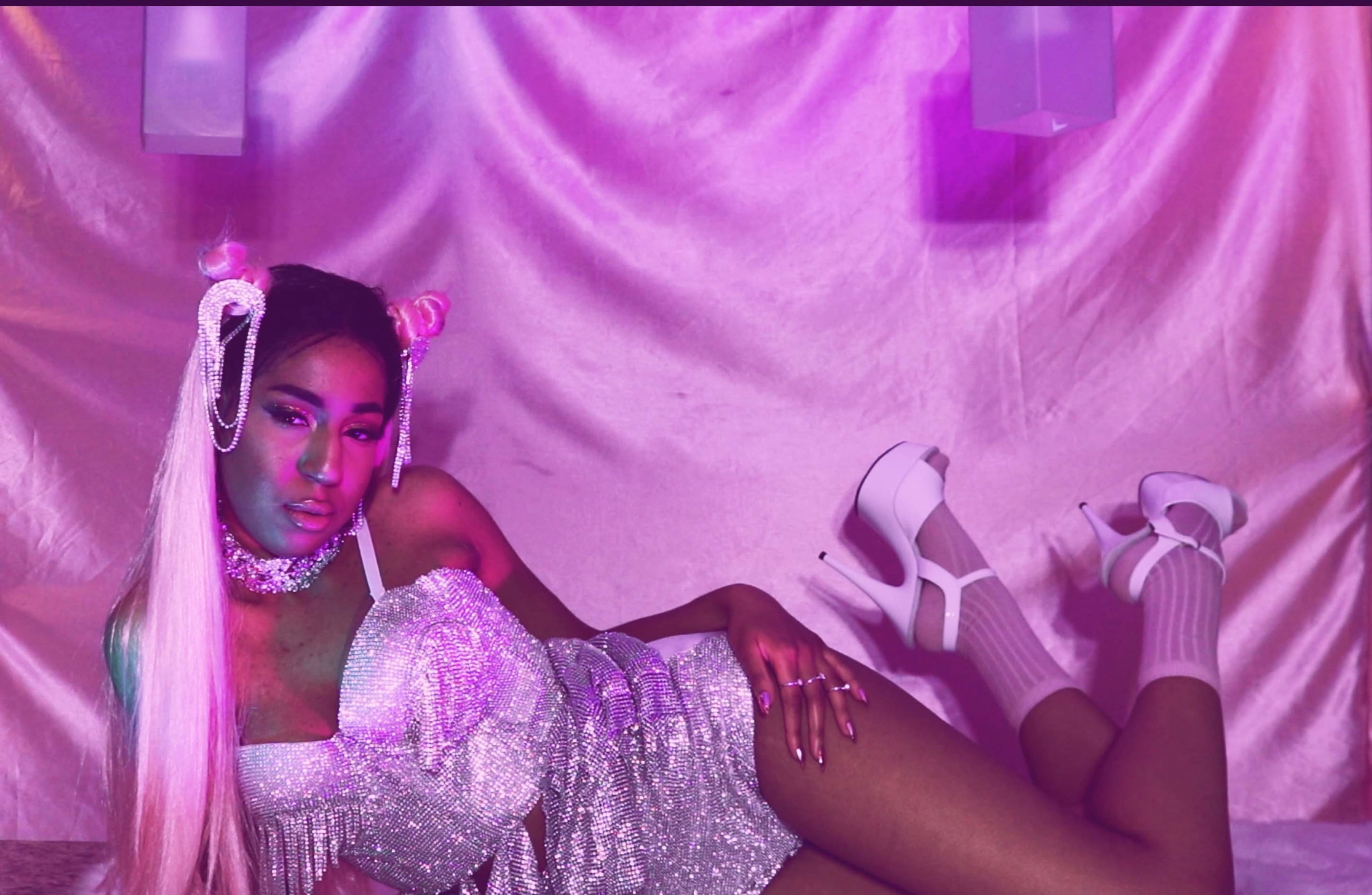 A person poses on a silky backdrop wearing a shimmering outfit and high heels, surrounded by soft purple lighting.