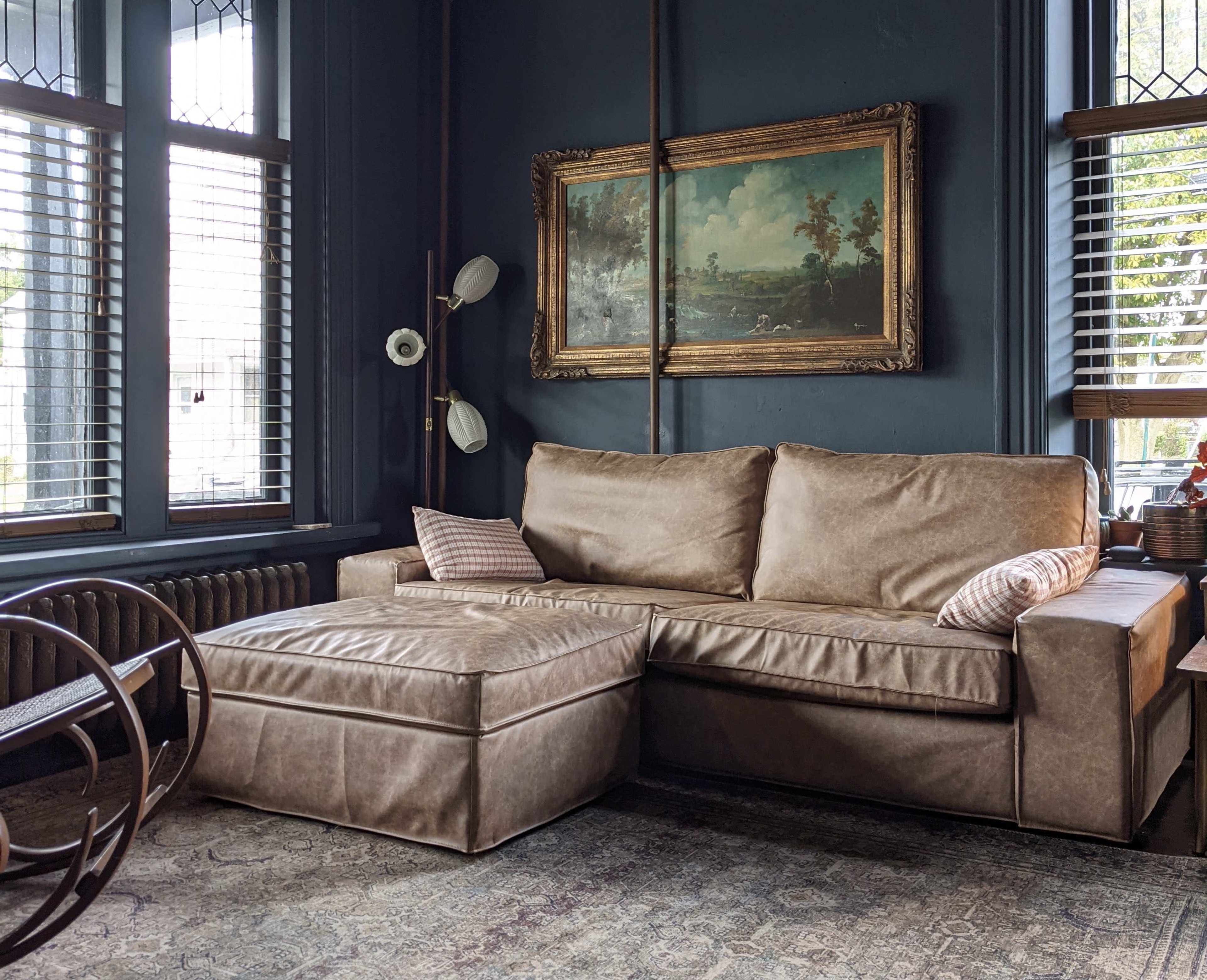 A modern, brown leather sectional sofa with an accompanying ottoman in a room featuring dark blue walls and large windows with blinds.