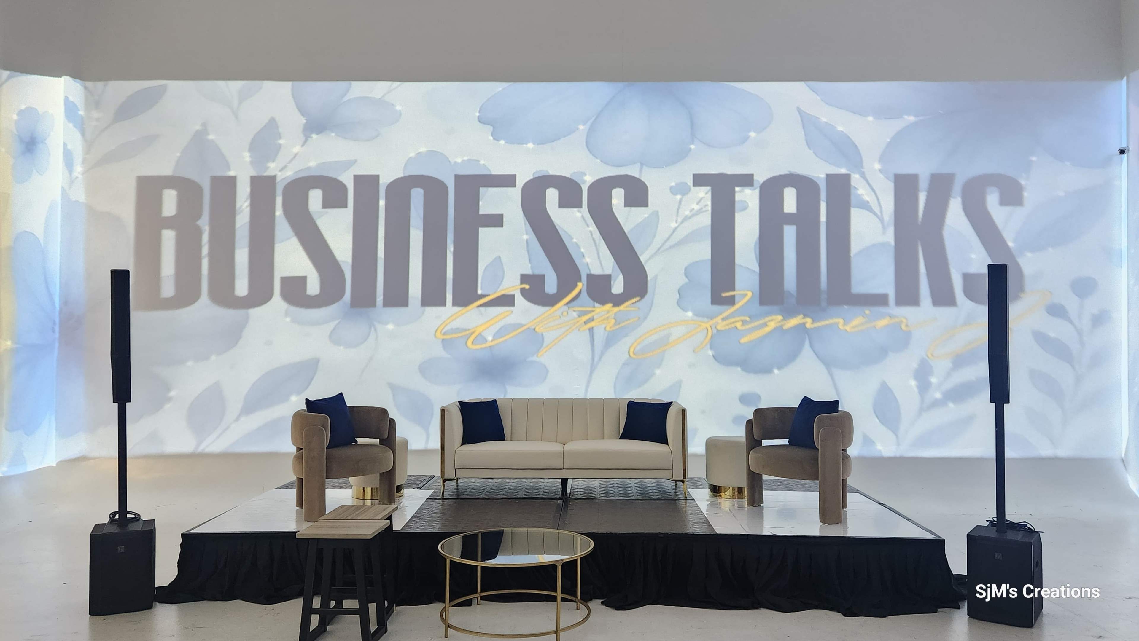 The image shows a stage setup for an event titled "Business Talks with Jasmine," featuring a light-colored backdrop with floral designs, a white couch, two armchairs, and a coffee table.