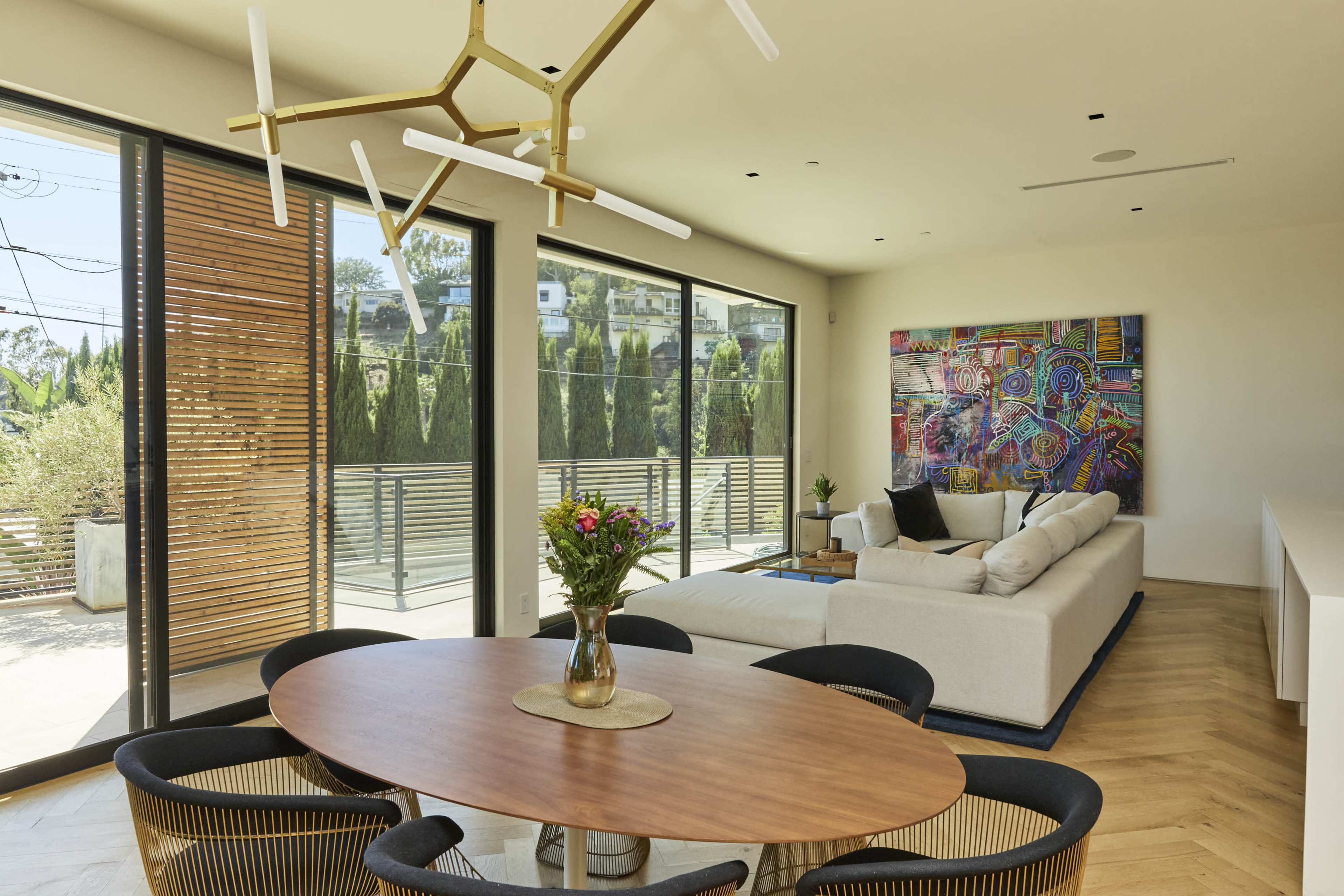 A modern living space features a round dining table with black chairs, a sectional sofa, colorful wall art, and large windows that provide views of an outdoor area.
