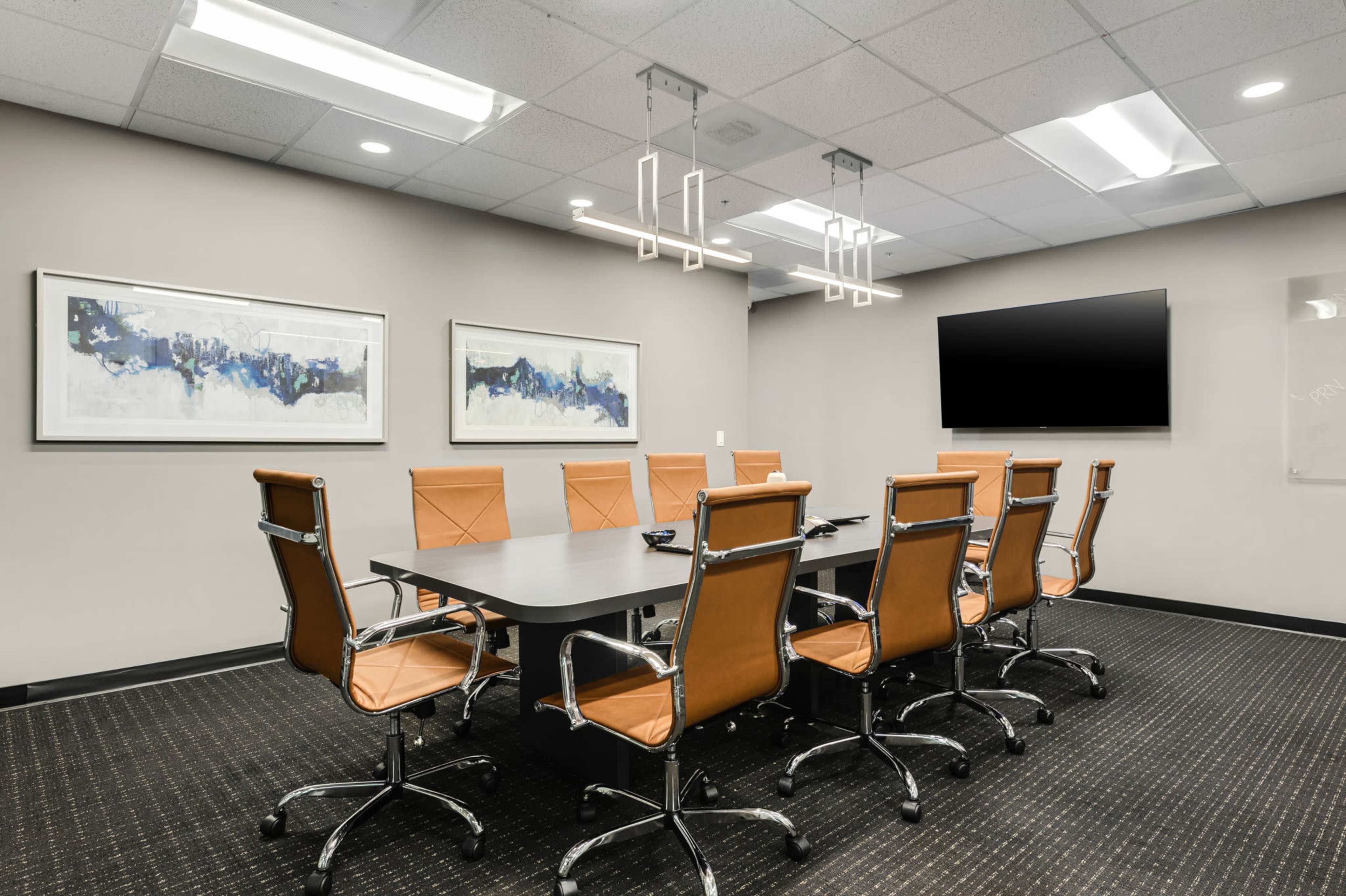 A conference room features a long table surrounded by eight brown leather chairs, with two abstract paintings on the wall and a wall-mounted television.