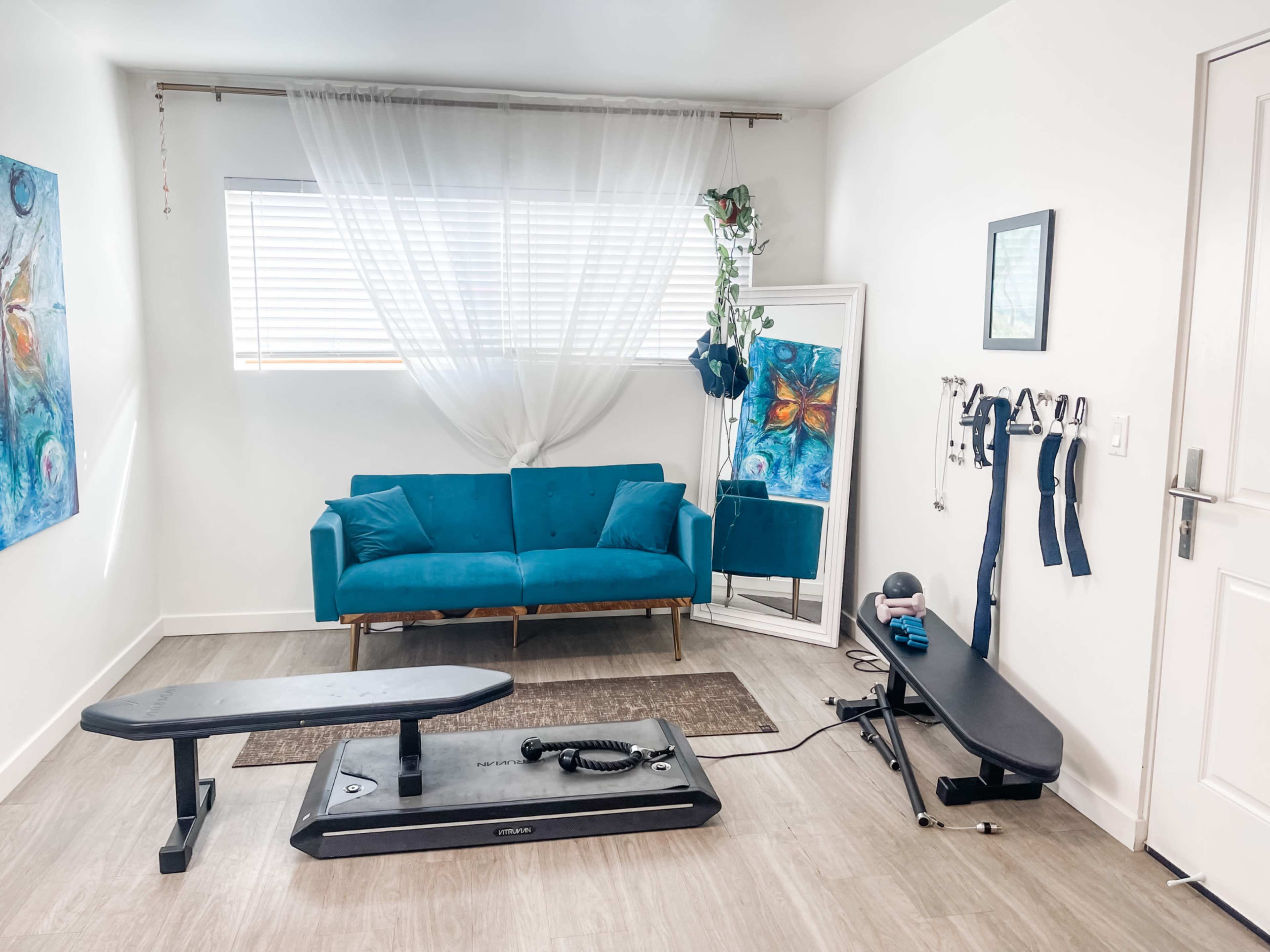 A fitness area features a blue couch, a mirror, and exercise equipment including a vibration plate and resistance bands against a white wall with a window.