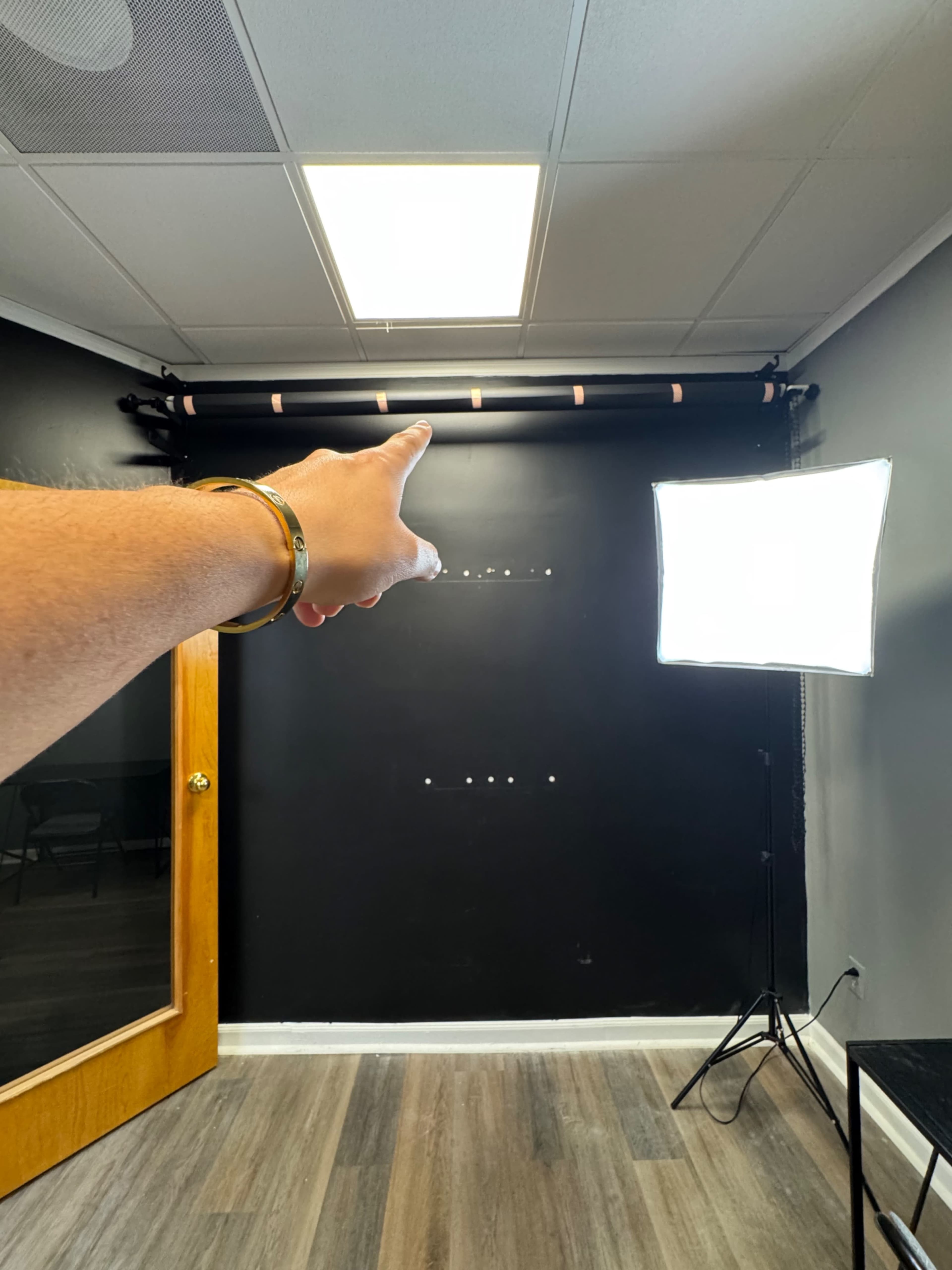 A person with a silver bracelet points towards a light fixture in a room with a dark wall and a softbox lighting setup.
