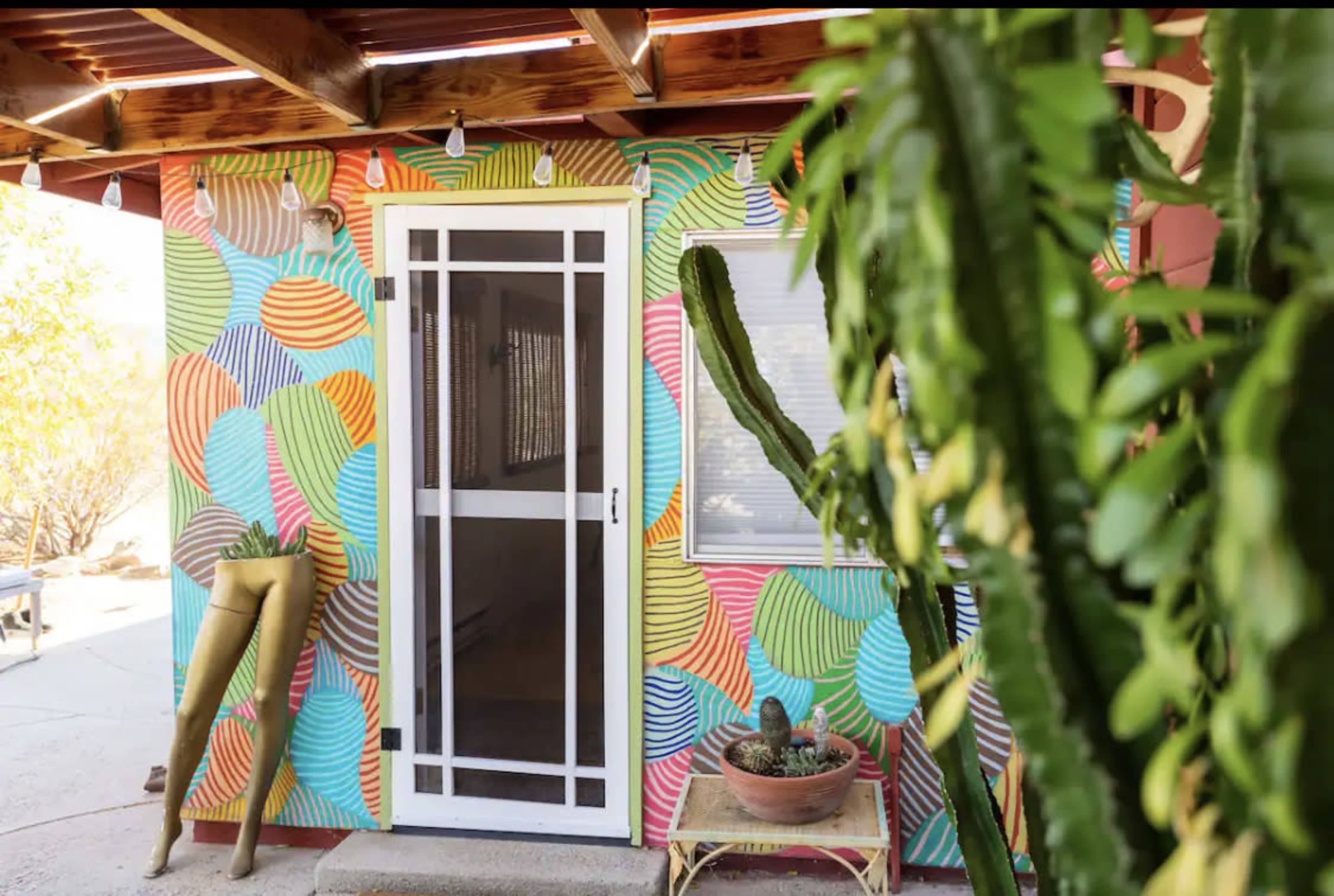 A brightly painted wall with colorful patterns surrounds a screened door, with a potted cactus and a mannequin leg visible in the foreground.