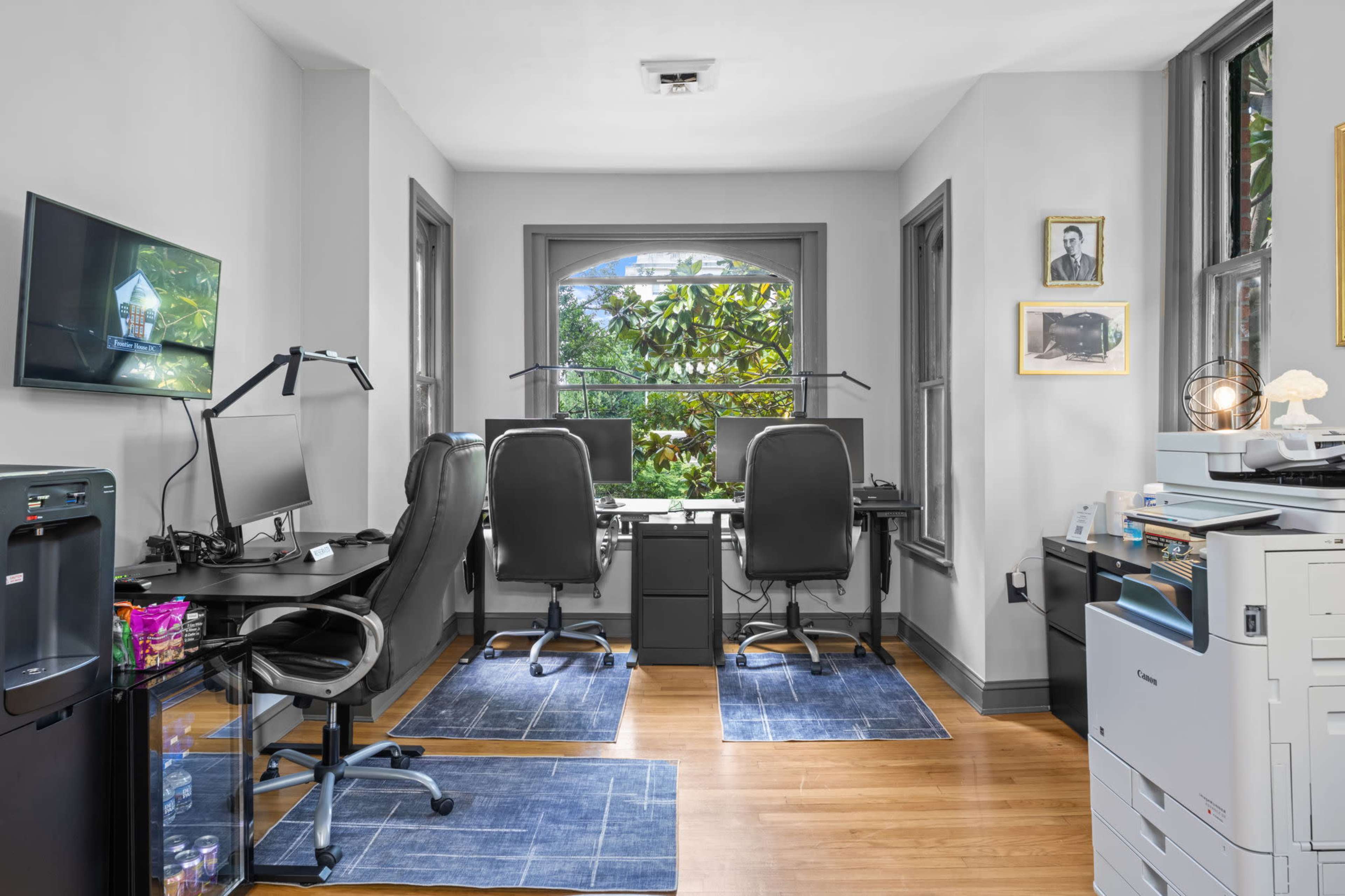 The image shows a modern home office with two black desks facing large windows, each equipped with computer monitors and stylish office chairs.