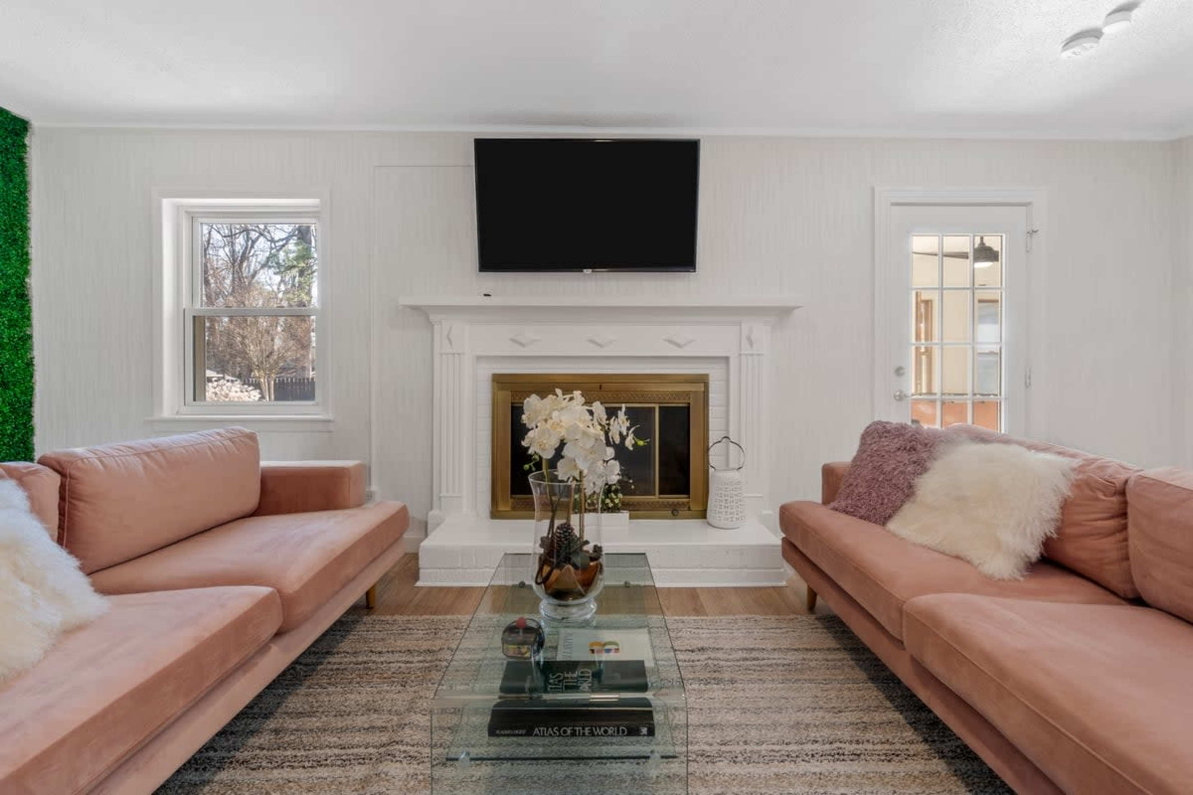 A living room features two pink sofas facing a glass coffee table, with a fireplace and a television mounted above it, flanked by windows.