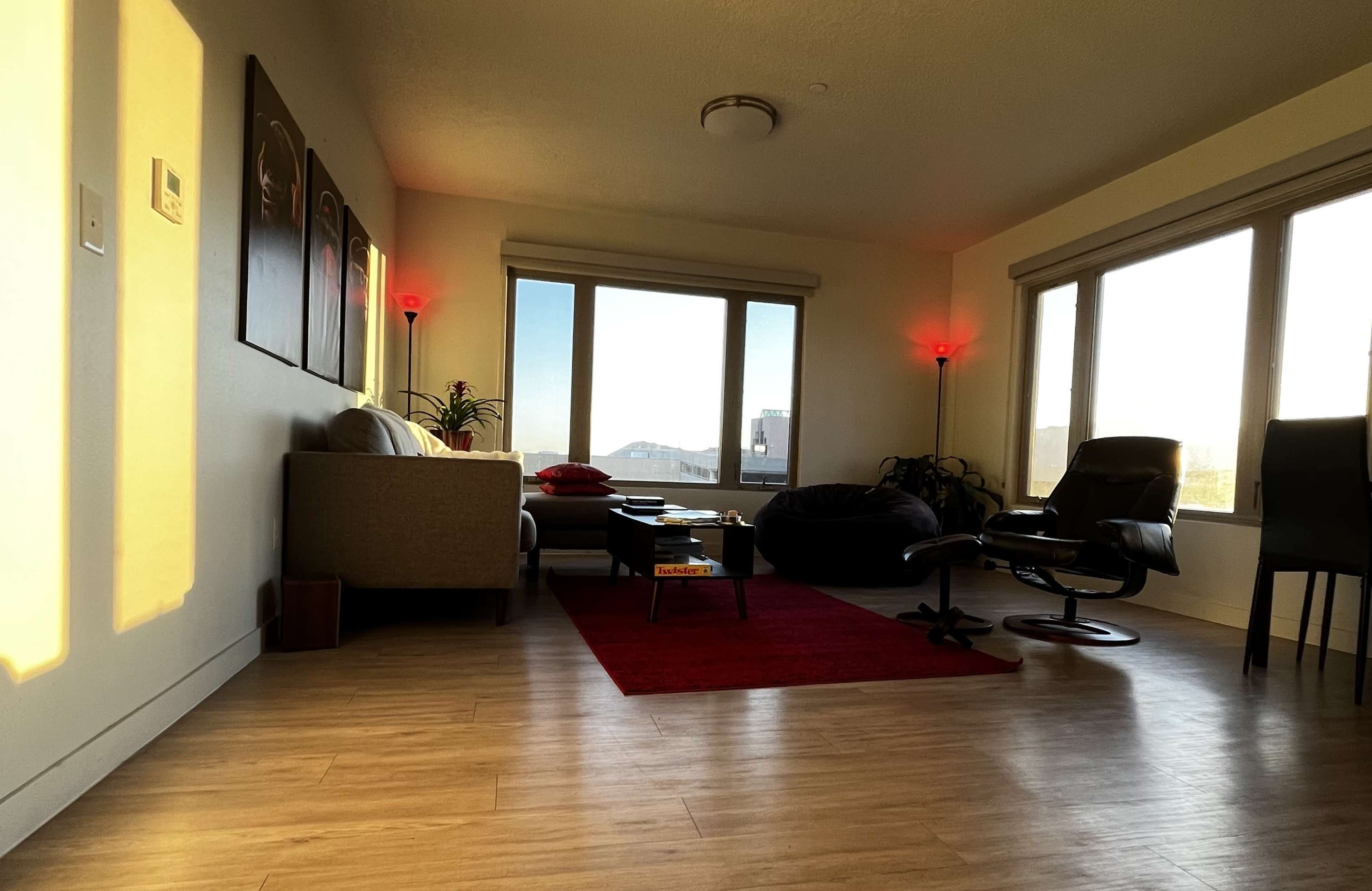 A modern living room with a sofa, a chair, and a red rug, illuminated by natural light from large windows.