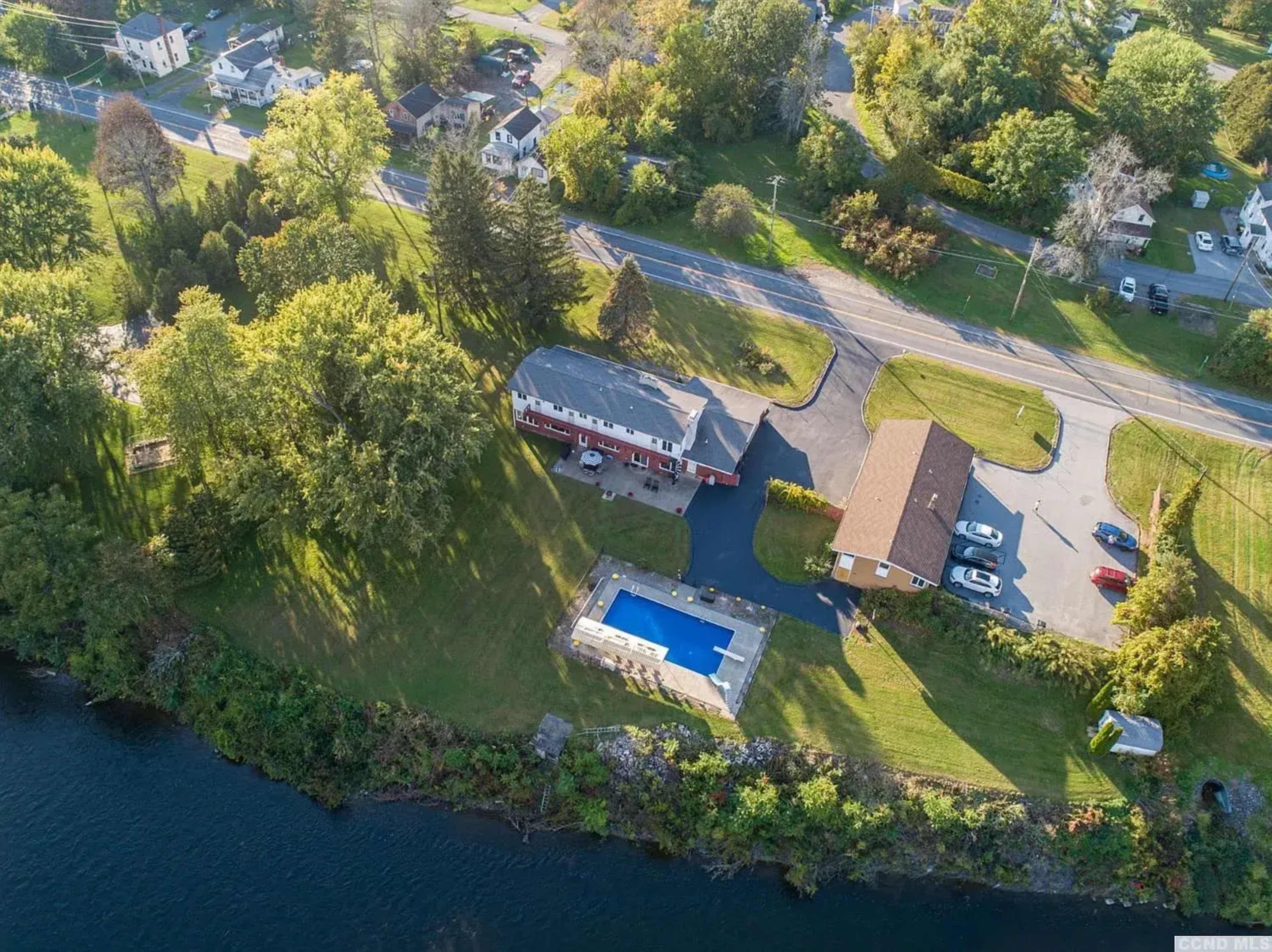 The image shows aerial view of a property with two buildings, a swimming pool, and surrounding greenery near a body of water.