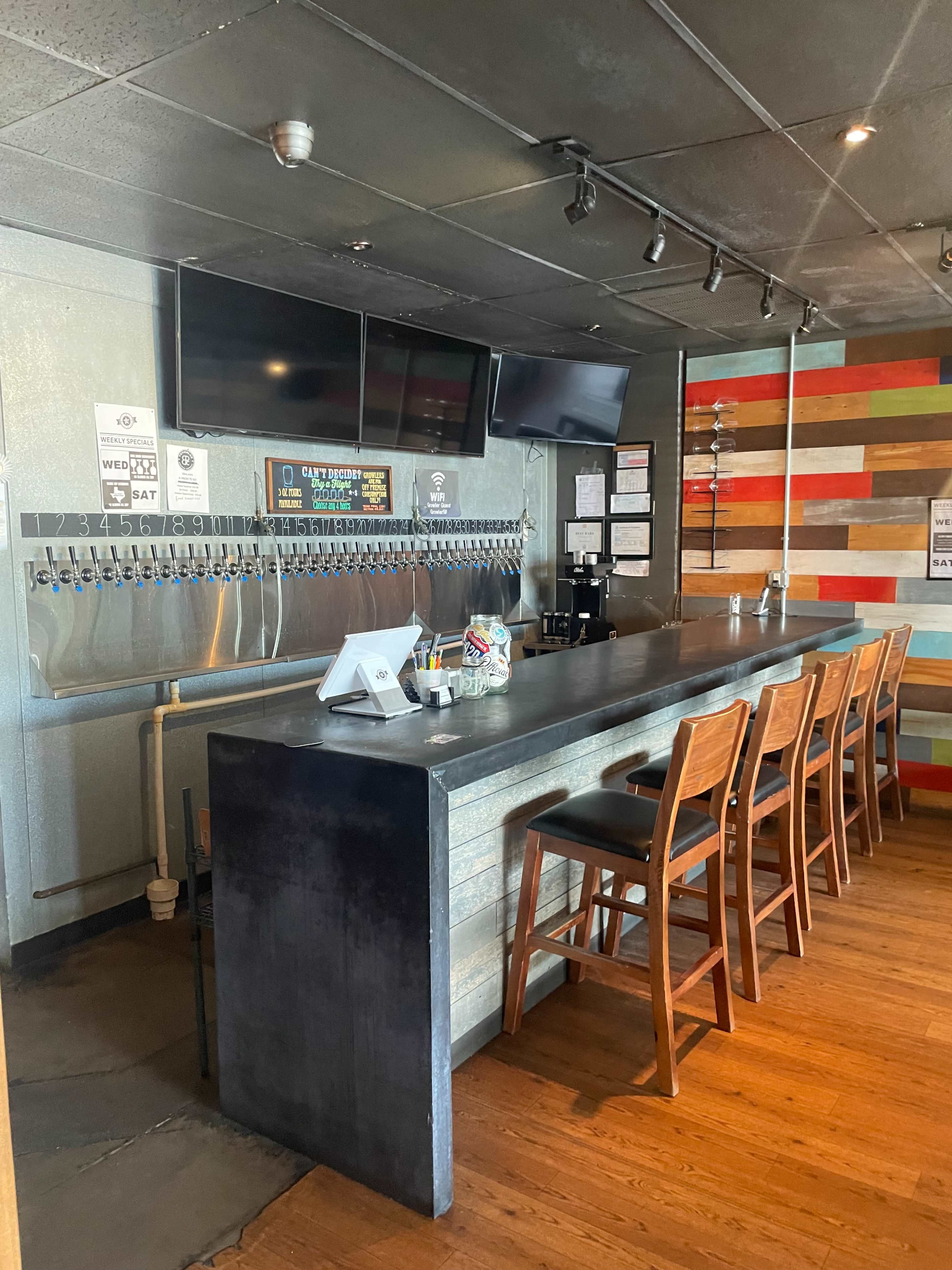 A modern bar with a long countertop, several rows of beer taps, and wooden stools against a colorful slatted wall.