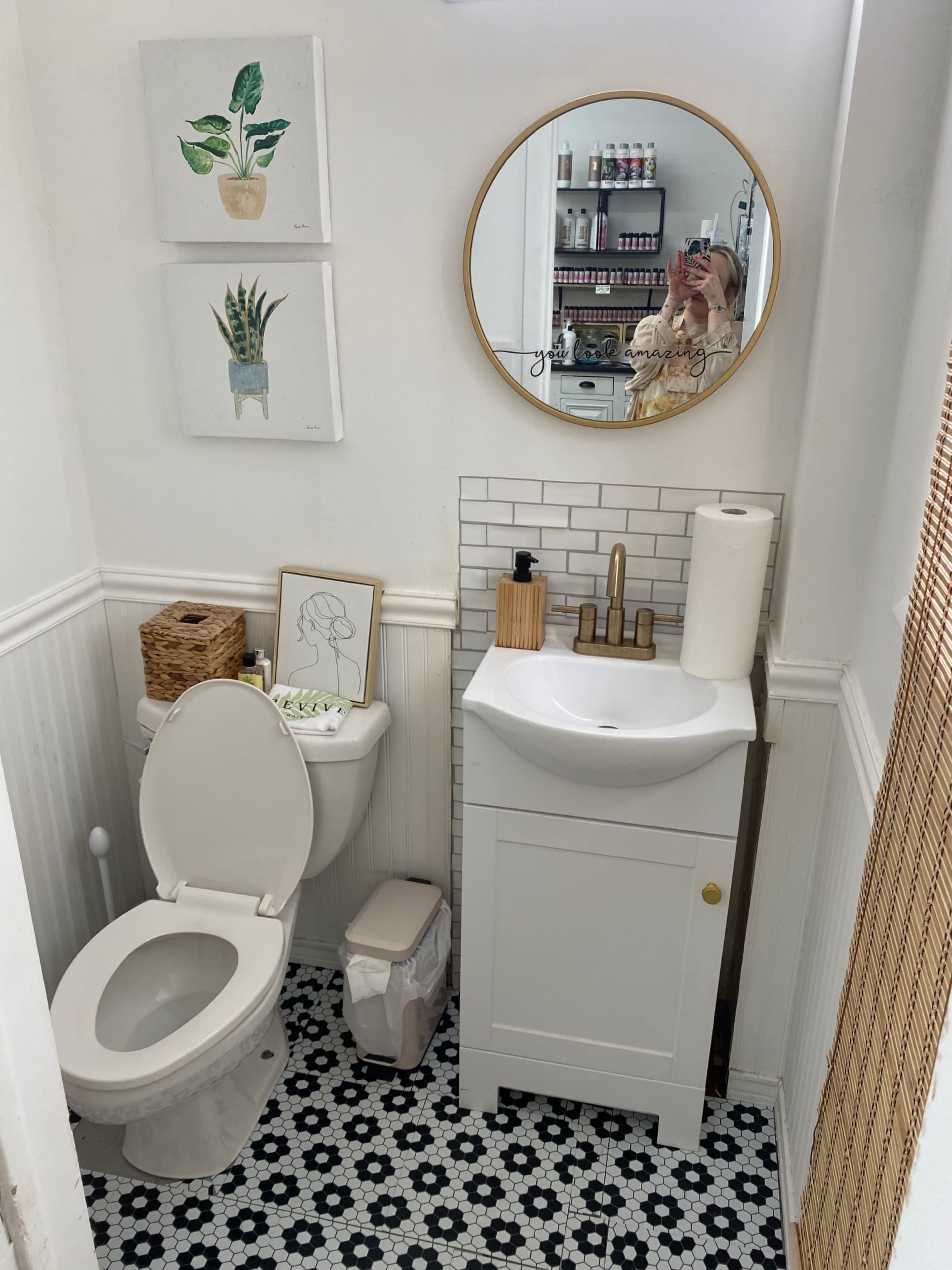 A small bathroom featuring a toilet next to a white vanity, a round mirror, and artwork of plants on the wall.