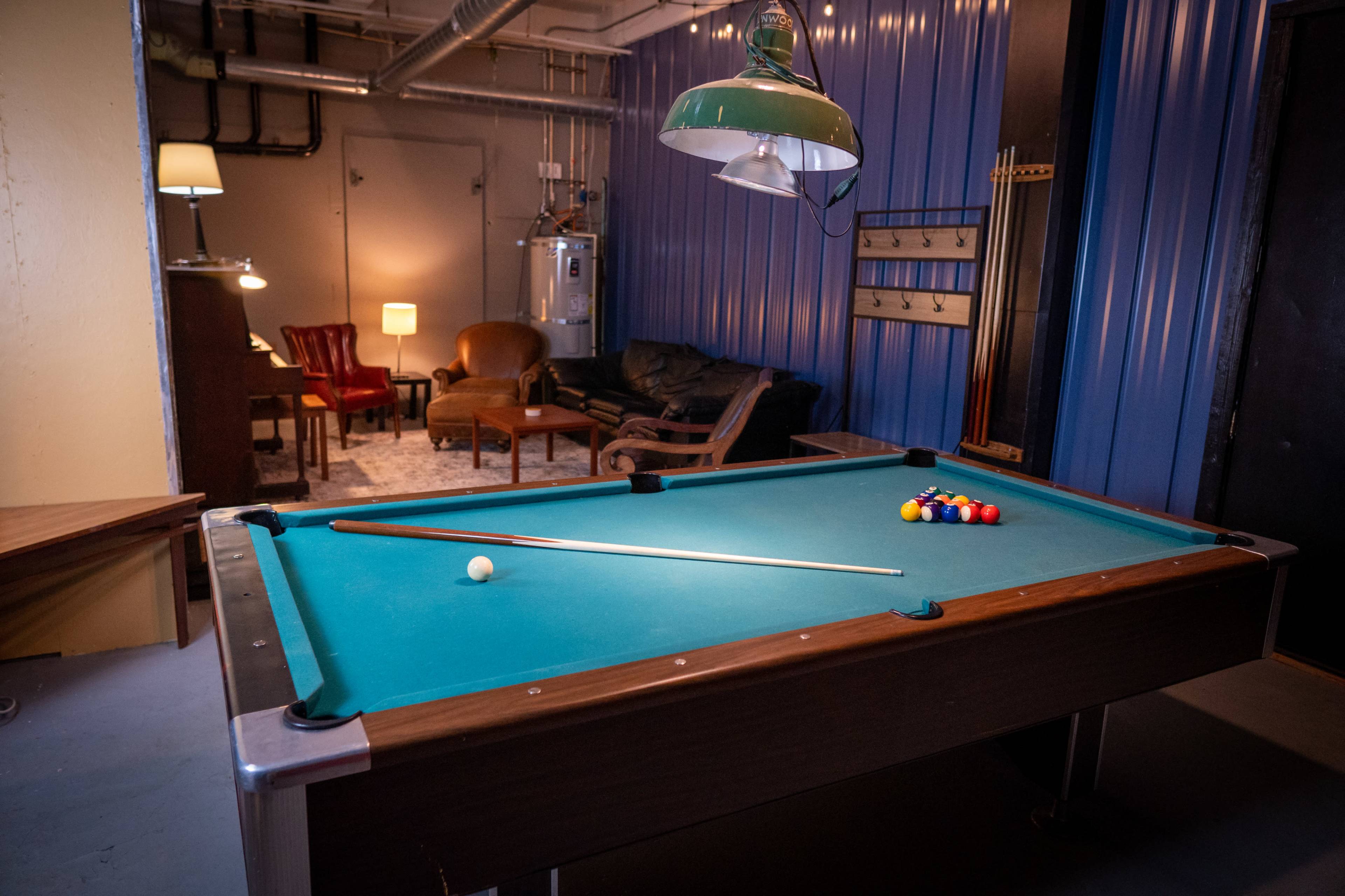 A well-lit room features a billiard table with a cue stick and balls arranged in a triangle, alongside a seating area with a couch and armchair.