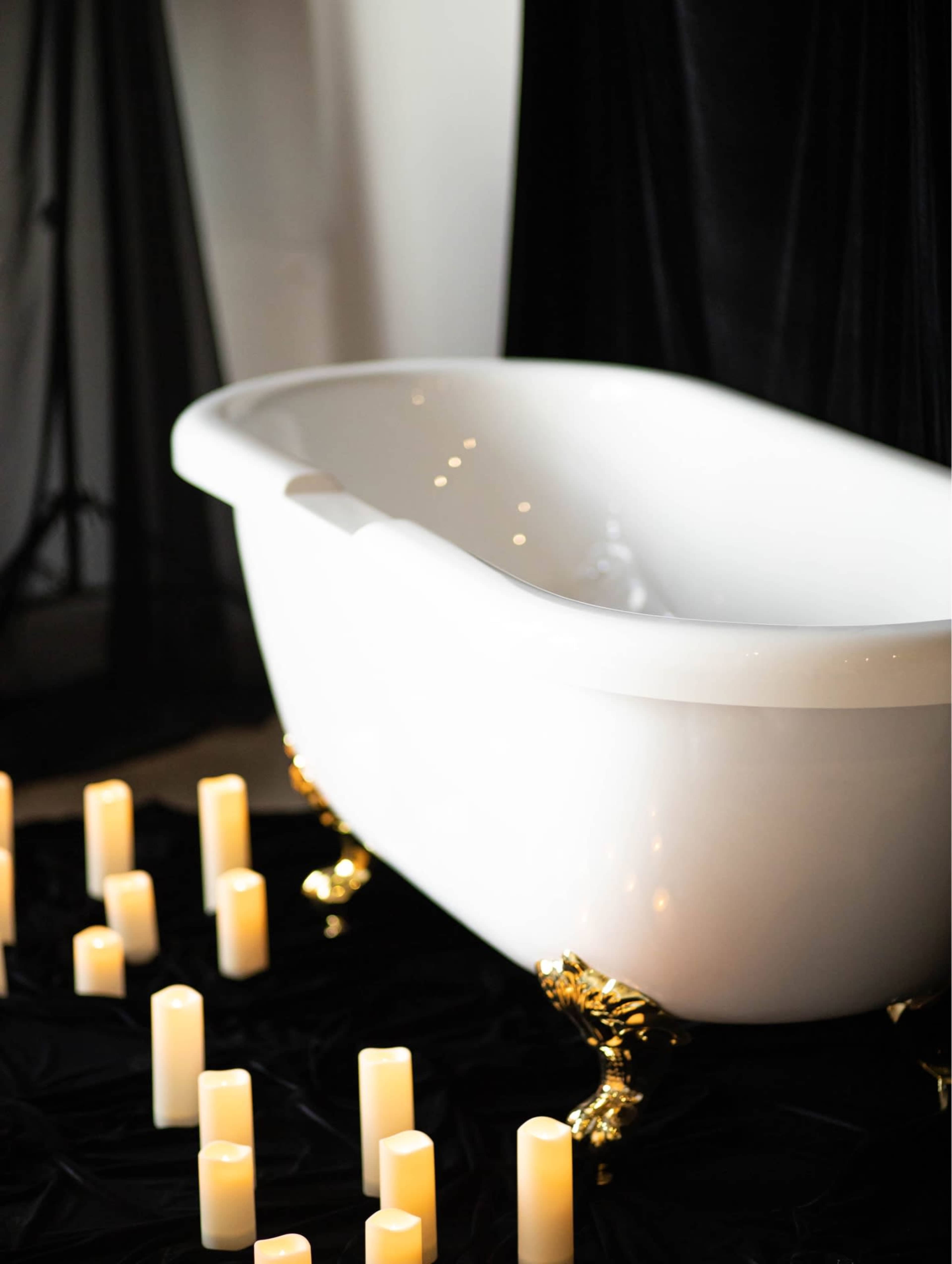 A white freestanding bathtub with gold feet is positioned on a dark surface surrounded by lit candles.