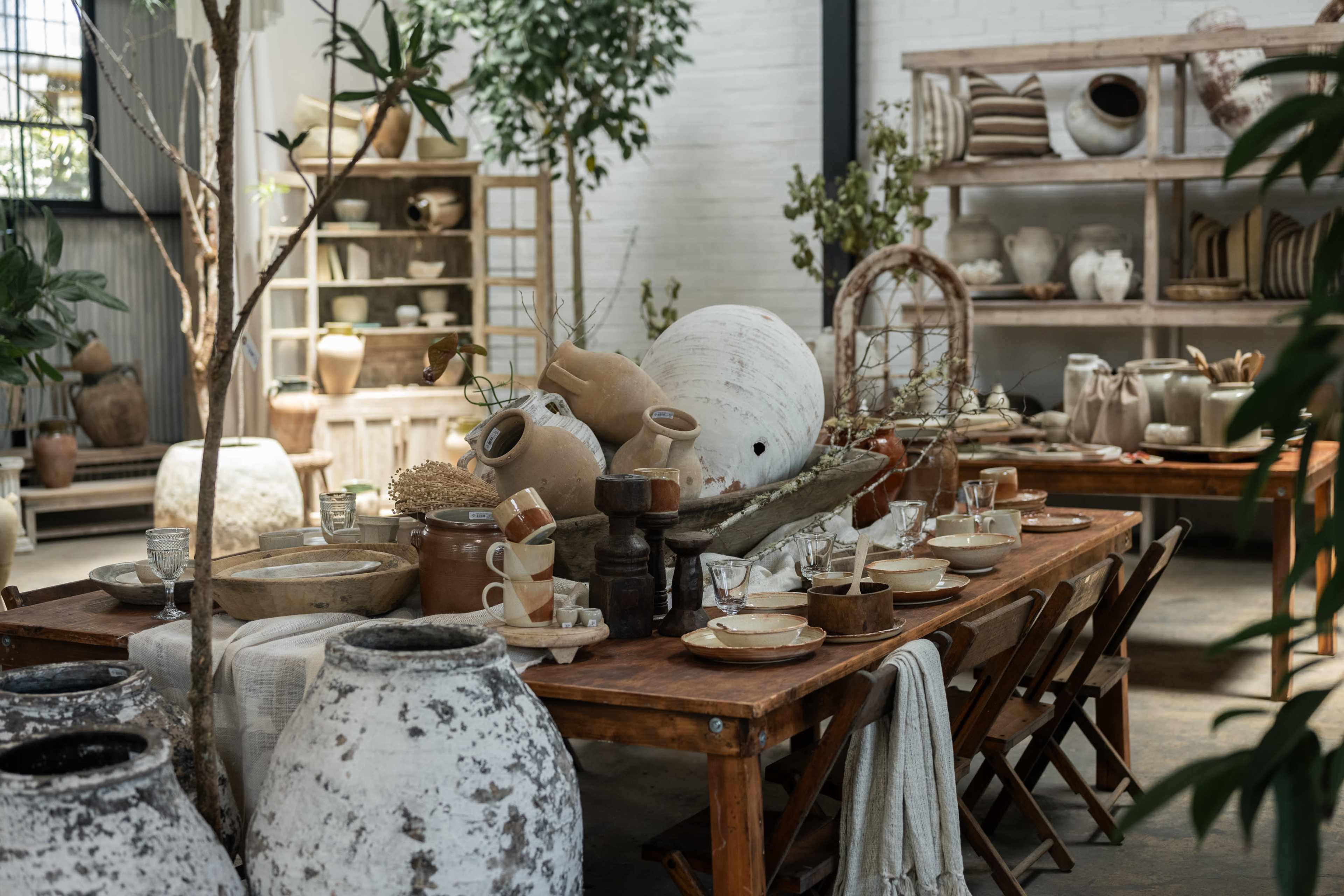 The image shows a rustic interior setting featuring a long wooden table set with dishes, surrounded by various pottery and decorative plants.