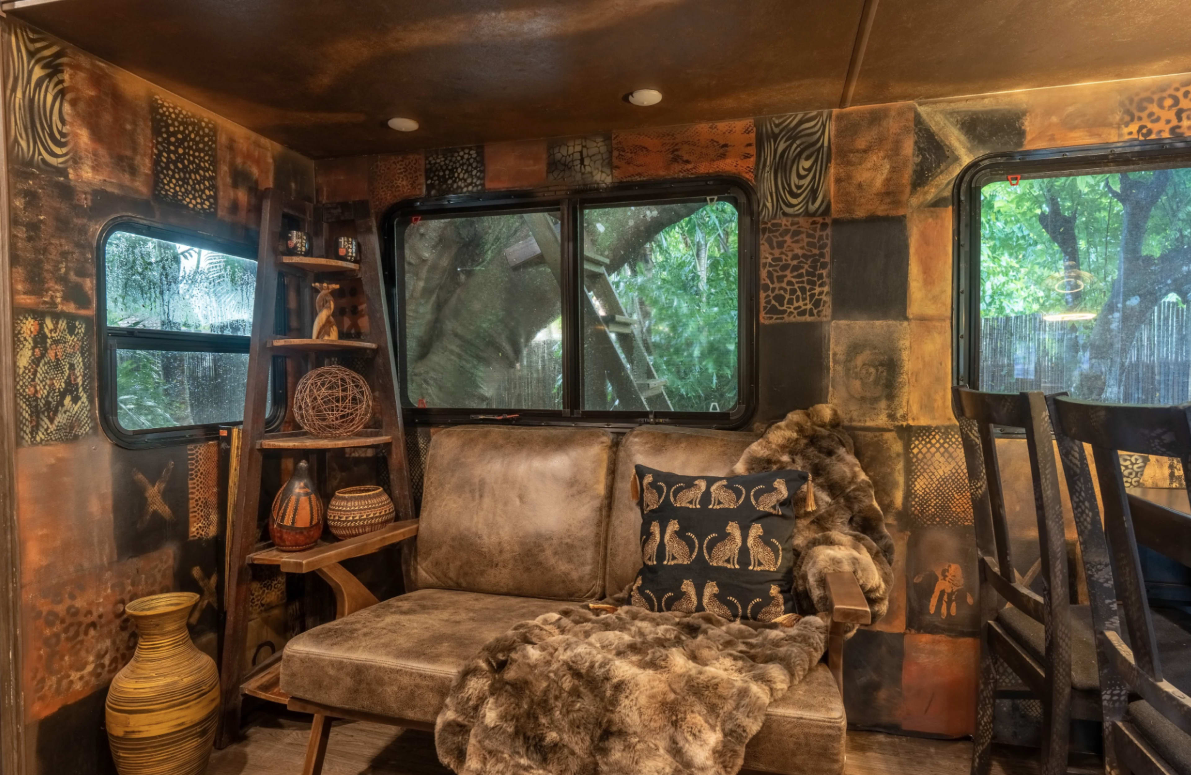The interior of a cozy RV features a brown sofa, a decorative fur throw, wooden shelves with items, and large windows that overlook greenery outside.