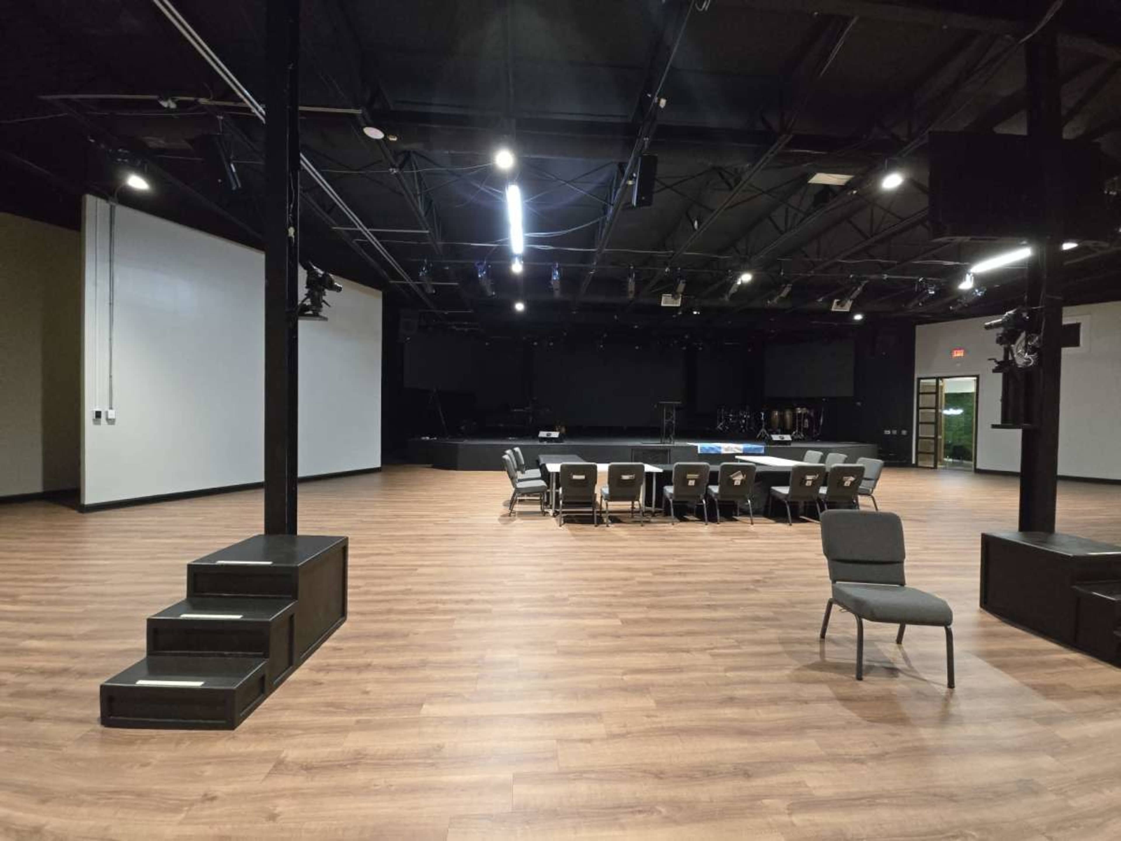 The image shows a spacious indoor venue with a wooden floor, featuring a central seating area arranged with chairs around a table, and a stage in the background.