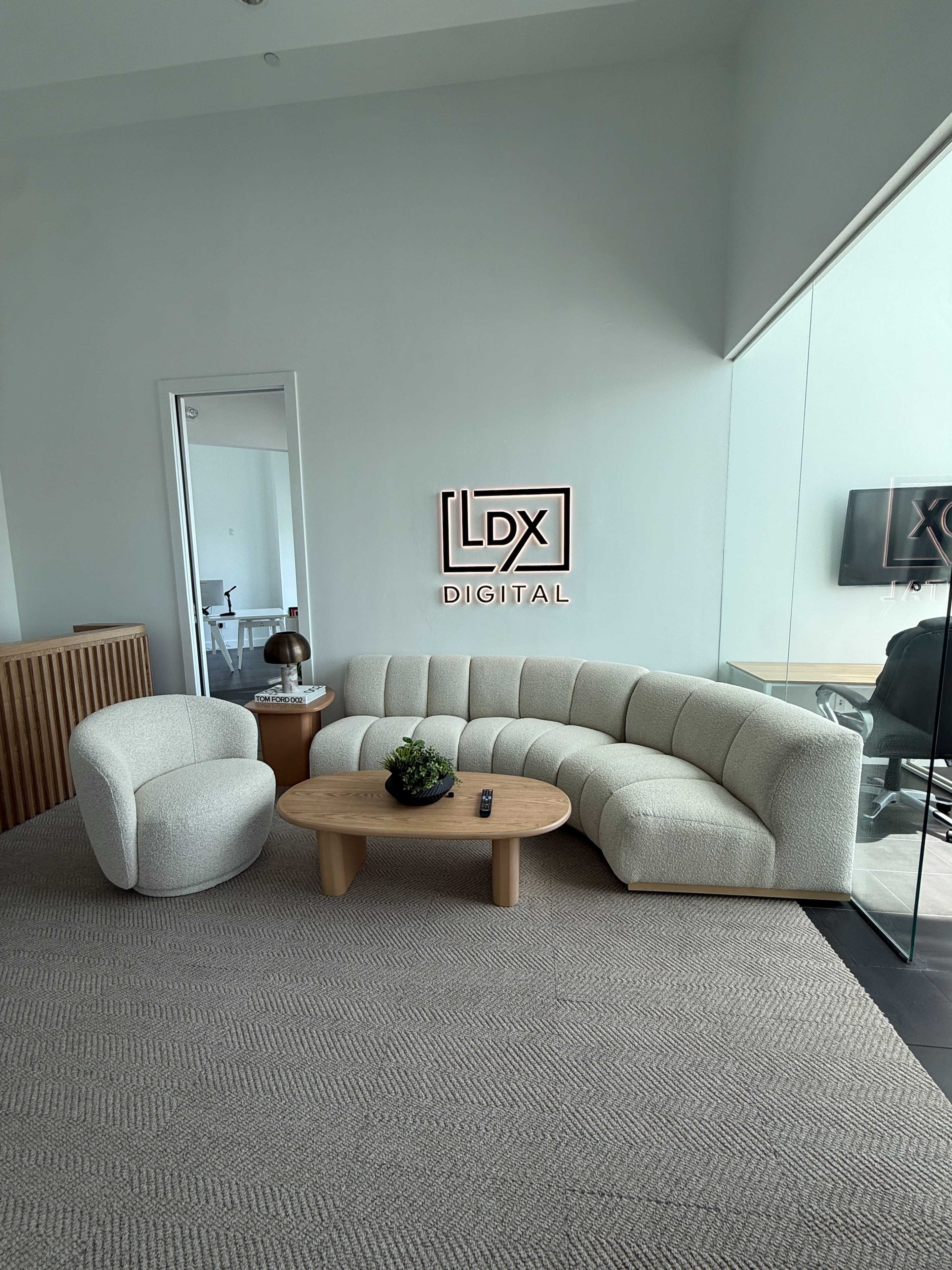 The image shows a modern lounge area featuring a curved white sofa, a round wooden coffee table, and a contemporary armchair, with a company logo on the wall.