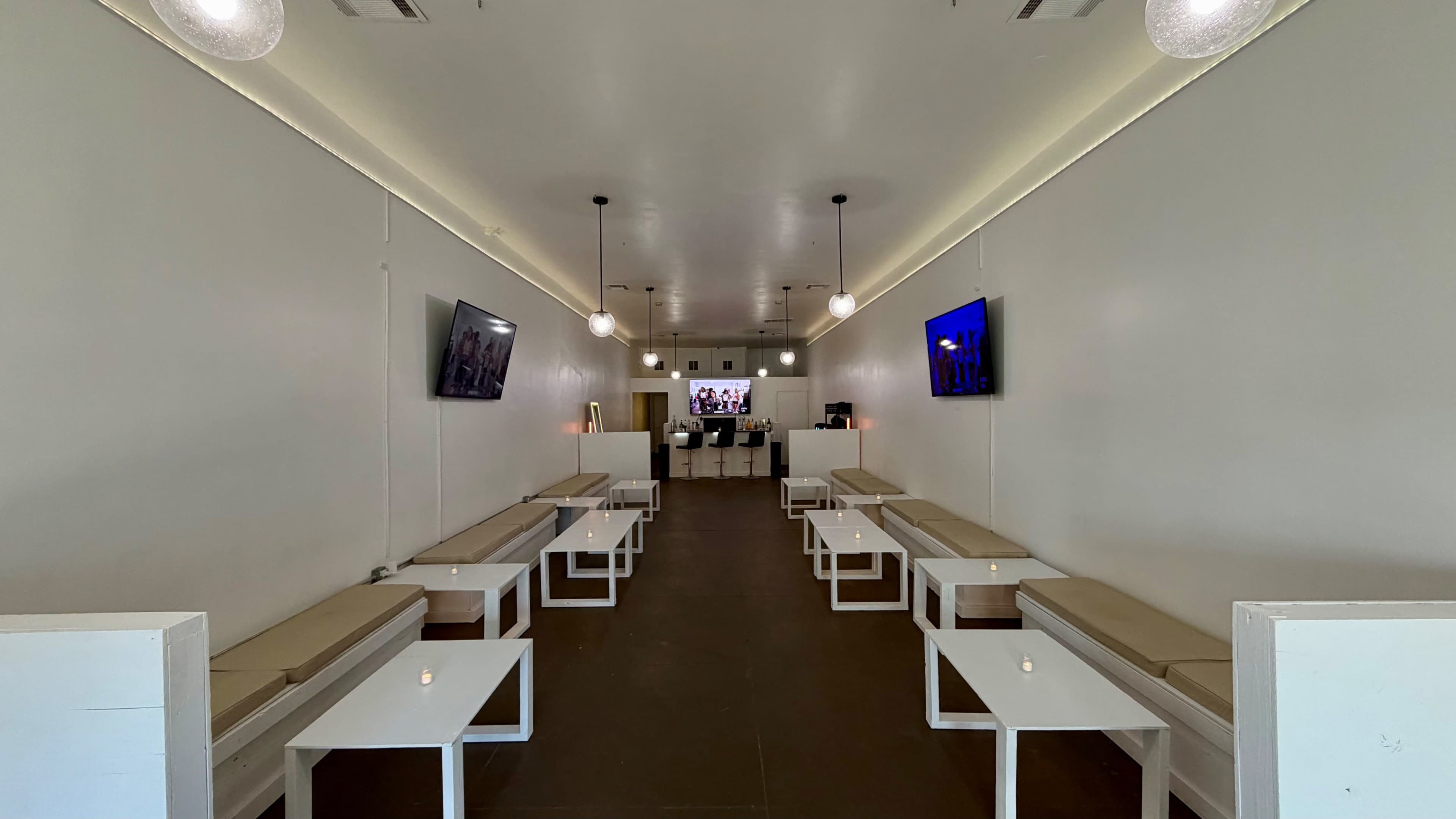 The image shows a modern, narrow lounge space with white benches, wall-mounted televisions, and soft overhead lighting.