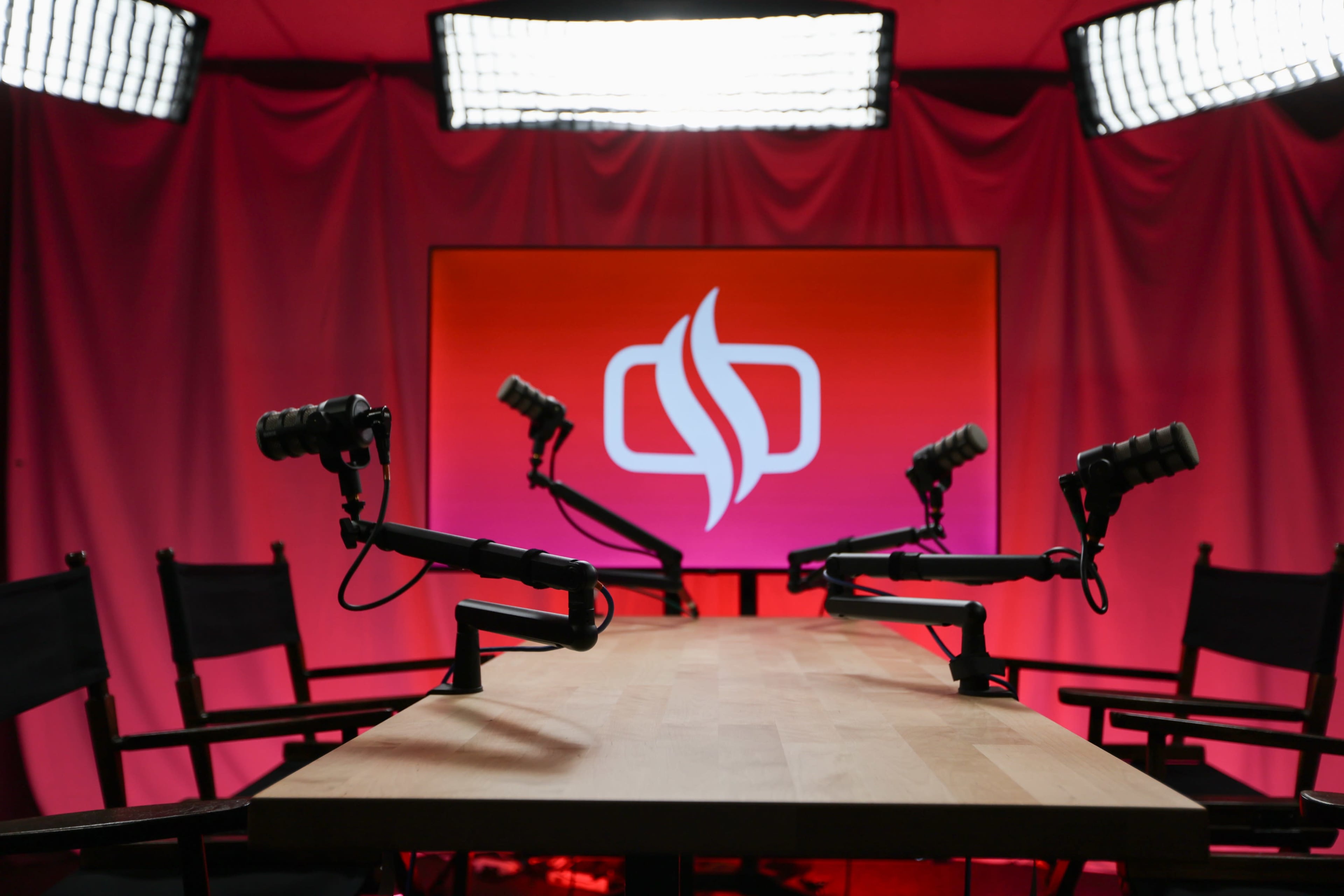 The image shows a podcast studio setup featuring a long wooden table, multiple microphone stands, and softbox lights illuminating a backdrop with a logo on it.