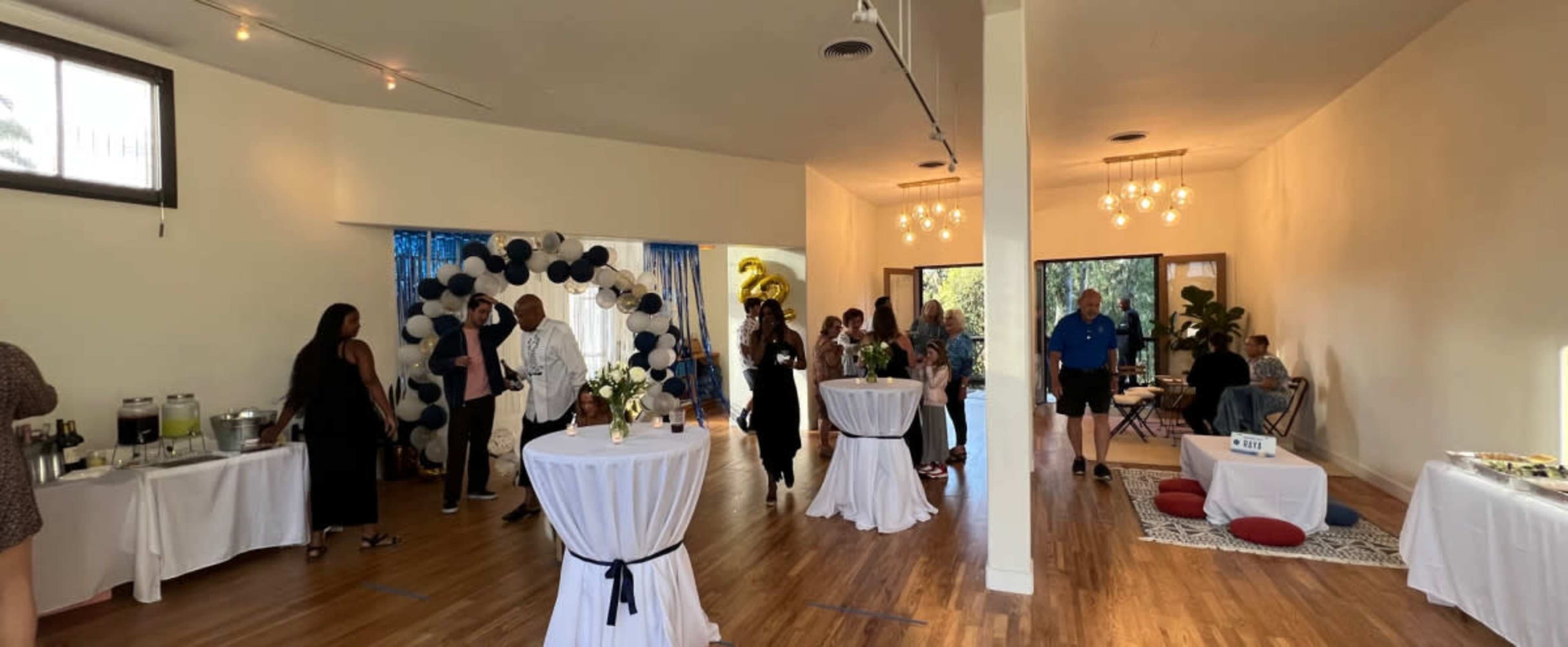 A group of people socializes at a spacious indoor event, featuring tables with refreshments and a decorative balloon arch in the background.