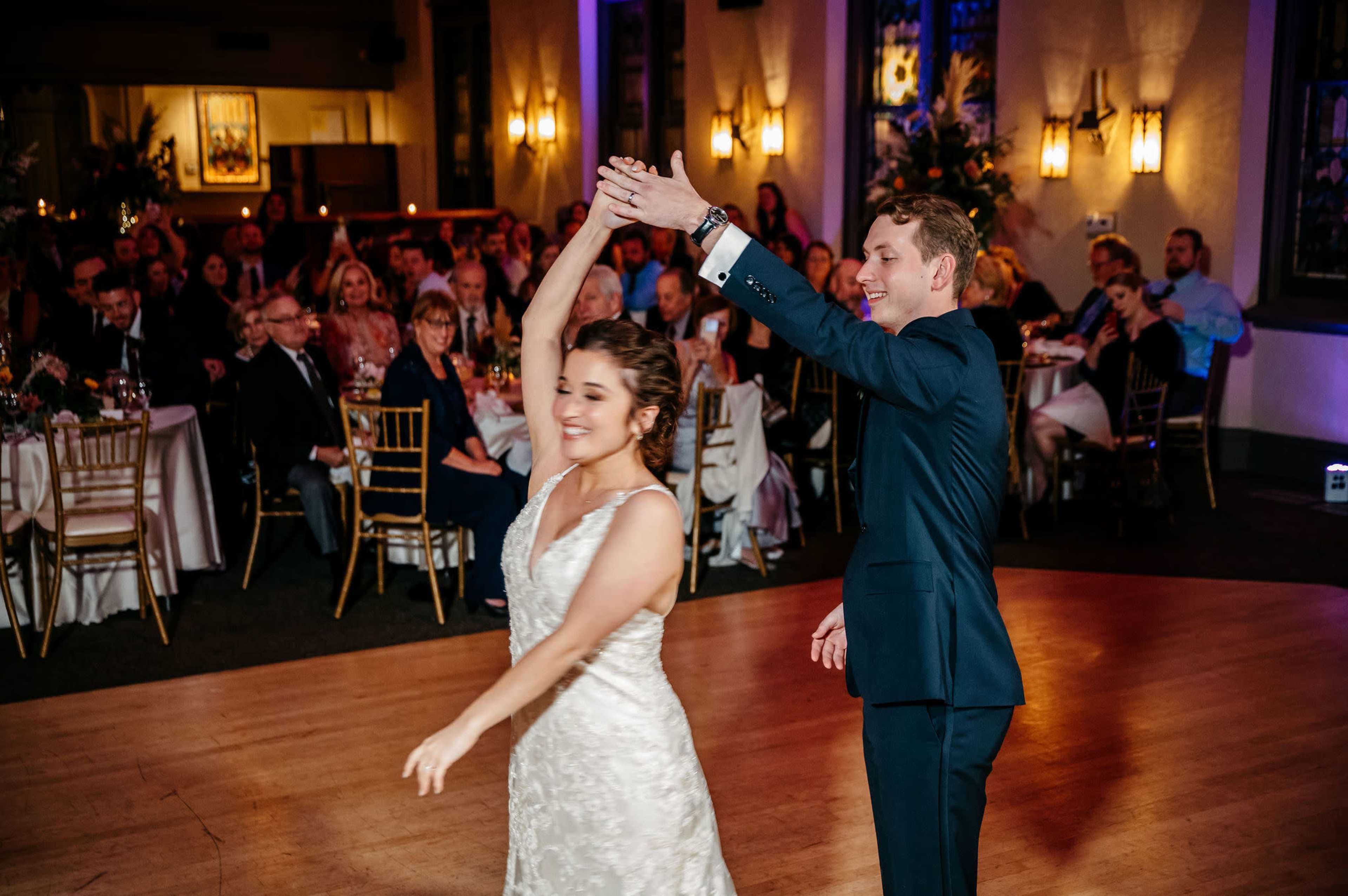 A couple dances together on a wooden floor in front of an audience at a reception venue.