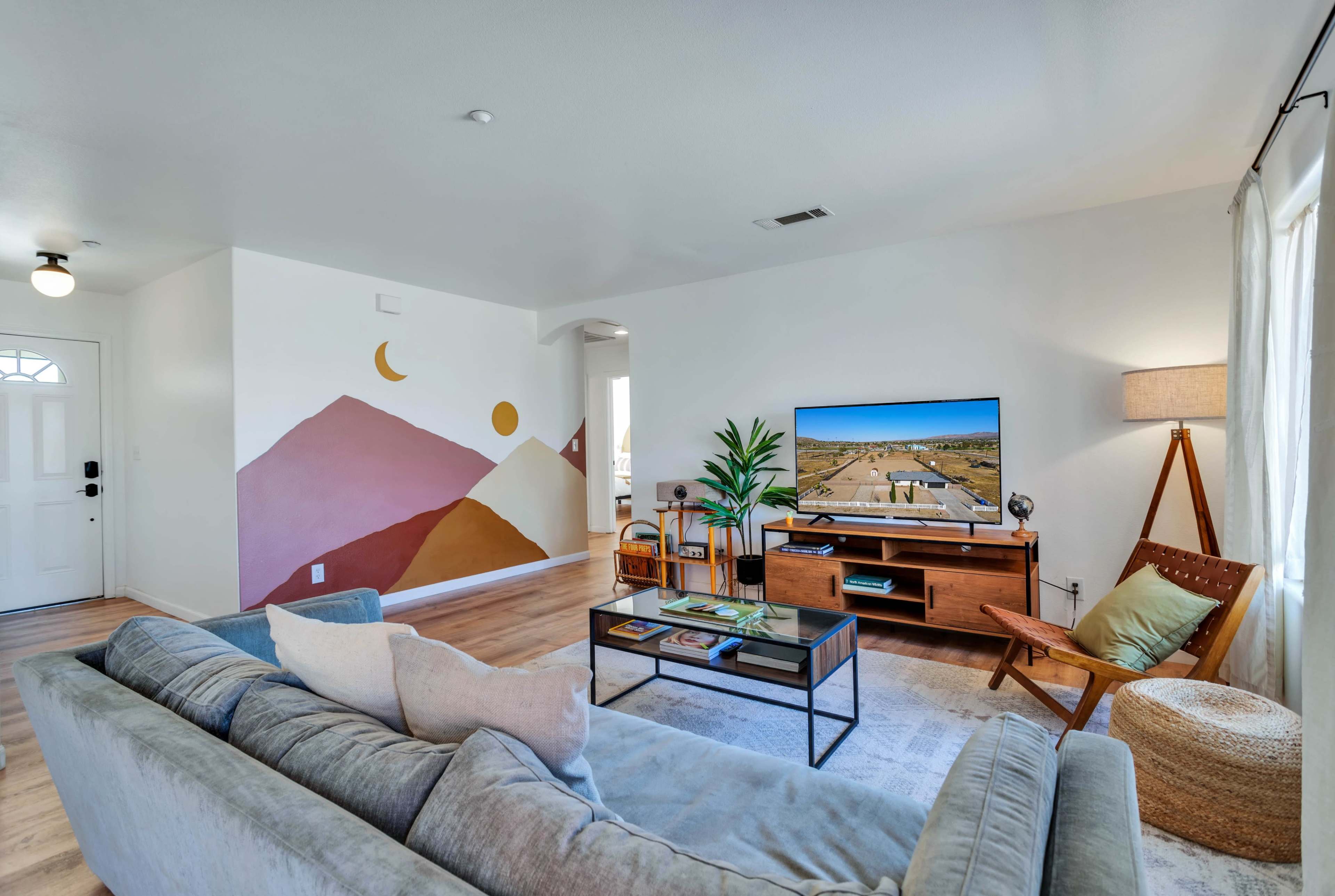 A living room features a modern couch, a television on a wooden console, and a mural of mountains and celestial shapes on one wall.
