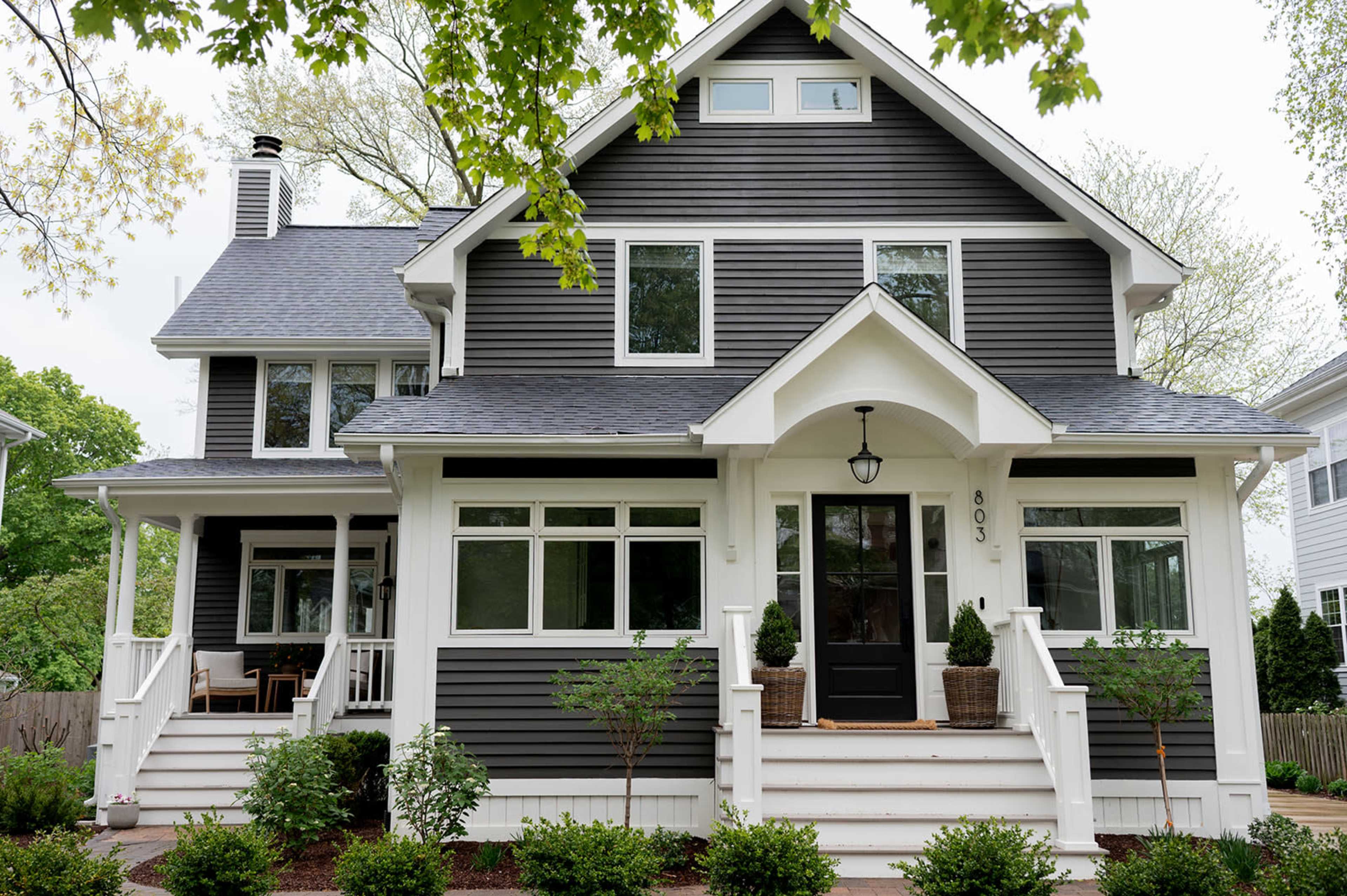 The image shows a large, two-story gray house with a steep roof, white trim, and a front porch flanked by neatly arranged shrubs.