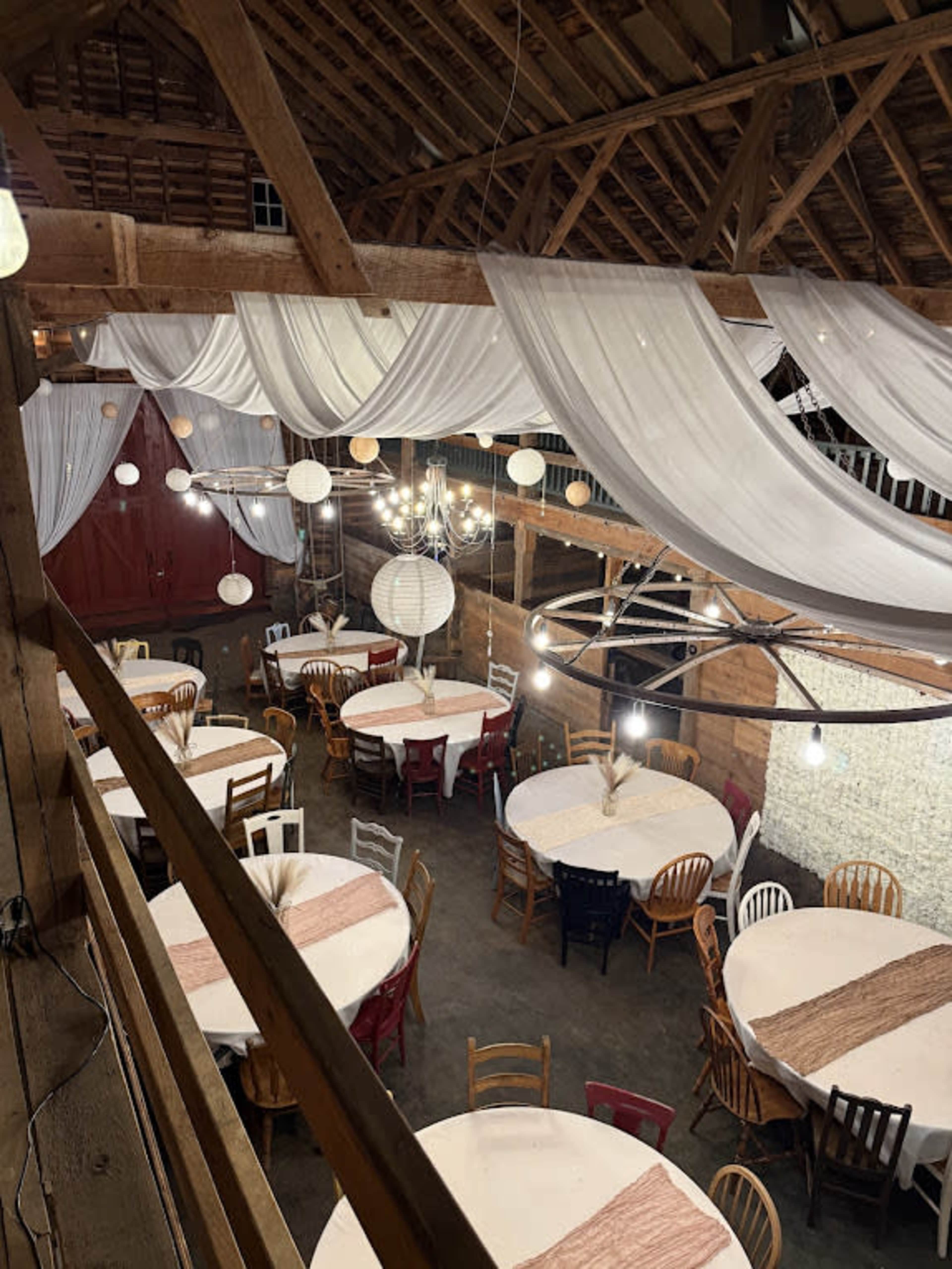 The image shows a rustic barn interior with multiple round tables draped in white tablecloths and decorated with hanging lights and draped fabric overhead.