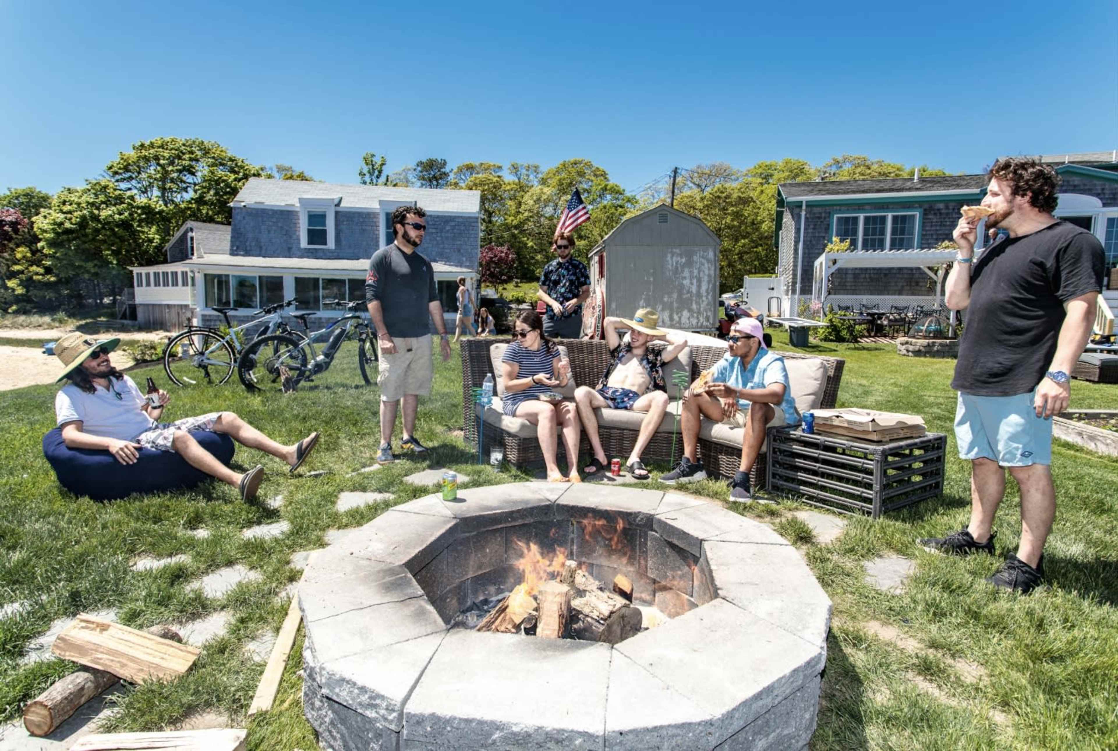 A group of people sits around a fire pit in a grassy area near several houses, with bicycles parked nearby.