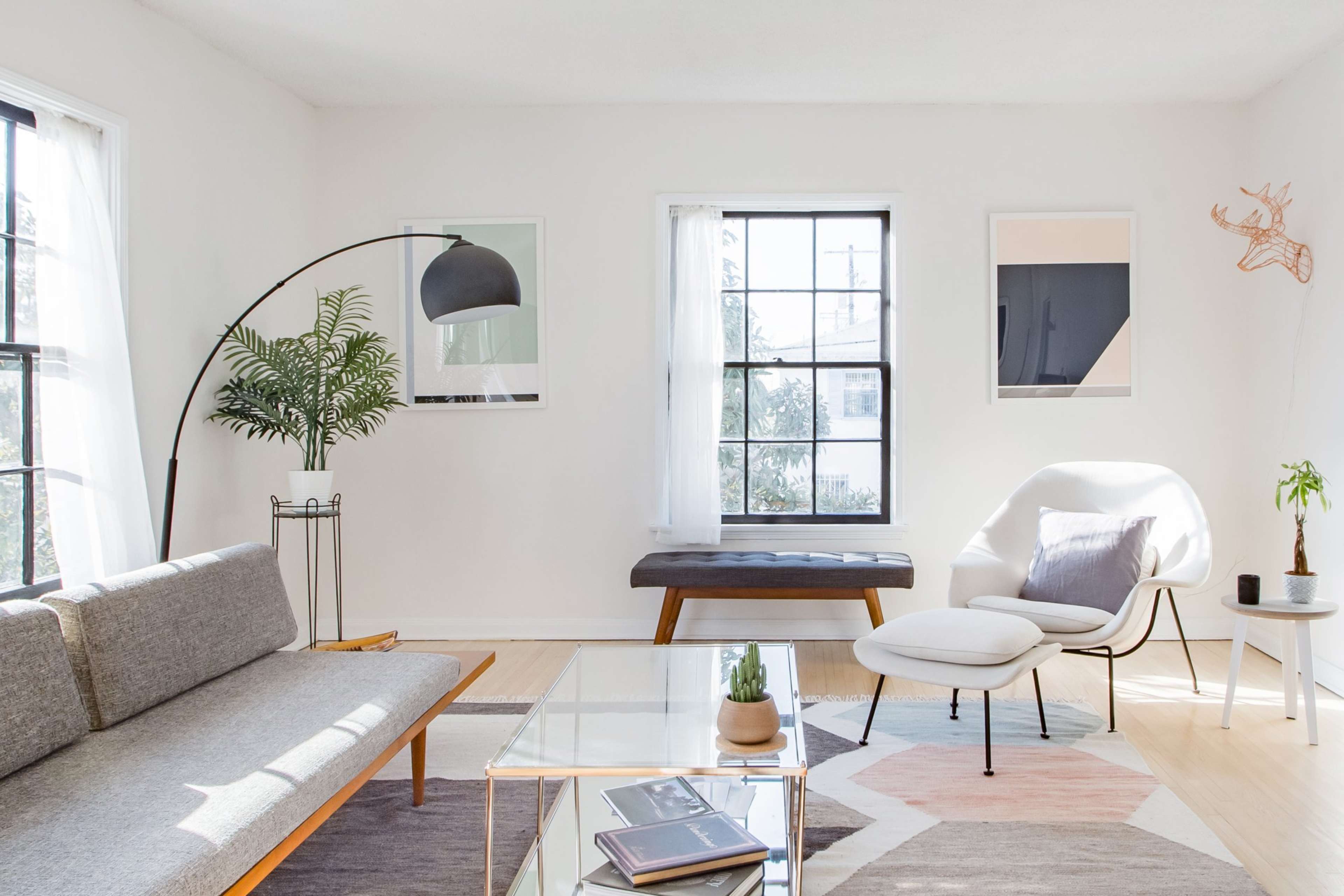 A modern living room features a gray sofa, a glass coffee table, a light chair, and large windows with a view of greenery outside.