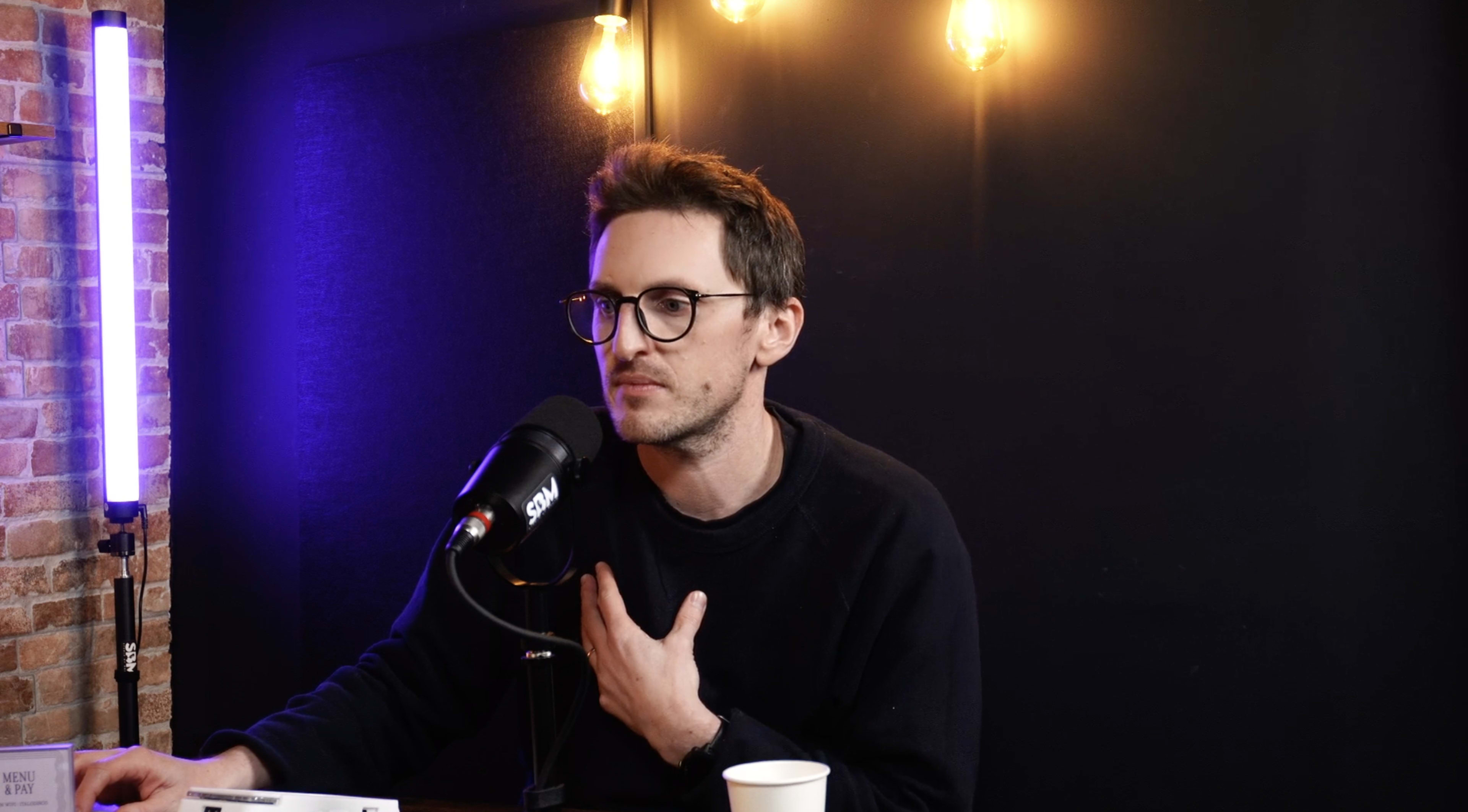 A person with glasses gestures while speaking into a microphone in a dimly lit room with exposed light bulbs.