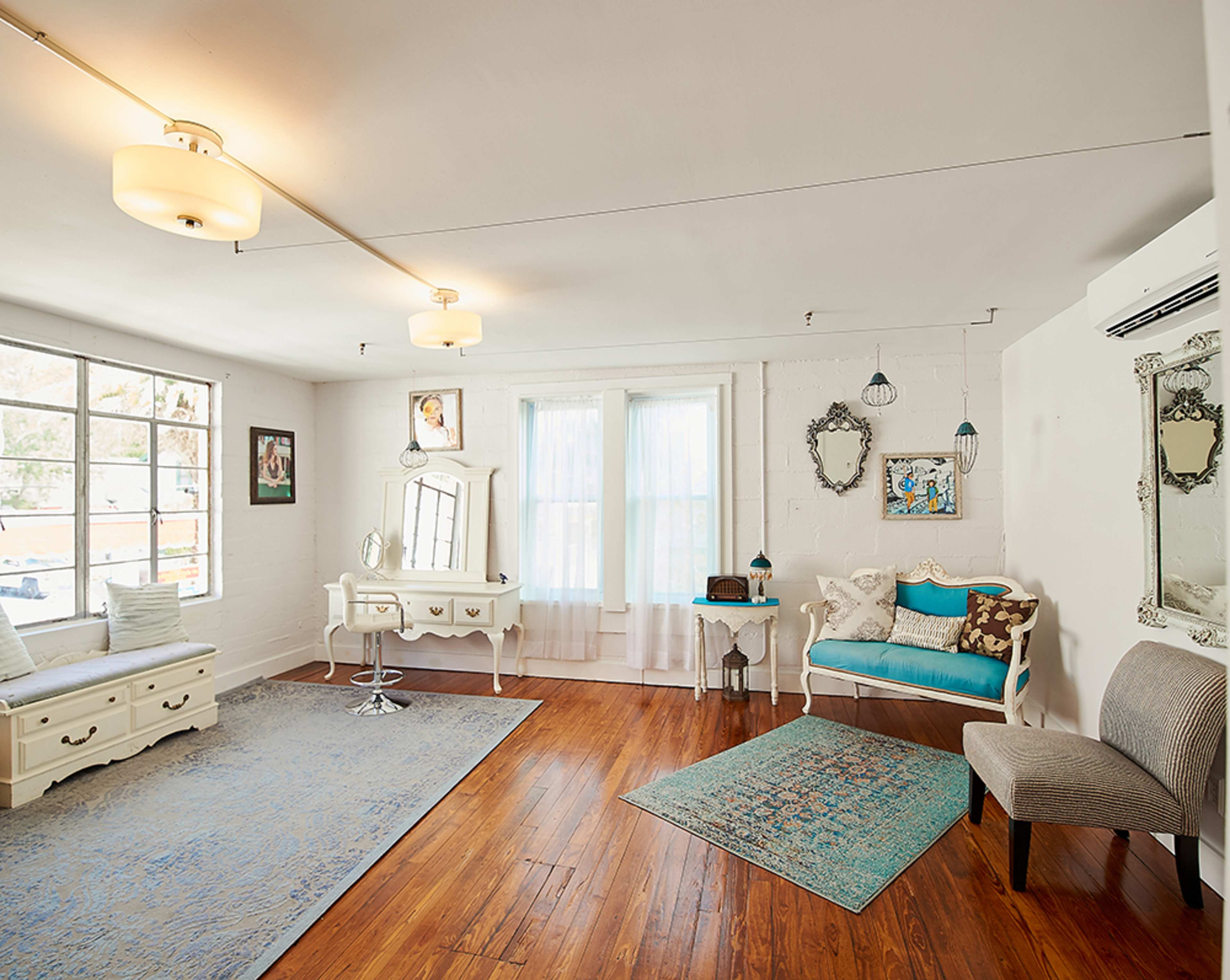 The image shows a bright, well-furnished room with wooden flooring, large windows, and various decorative mirrors and furniture pieces.