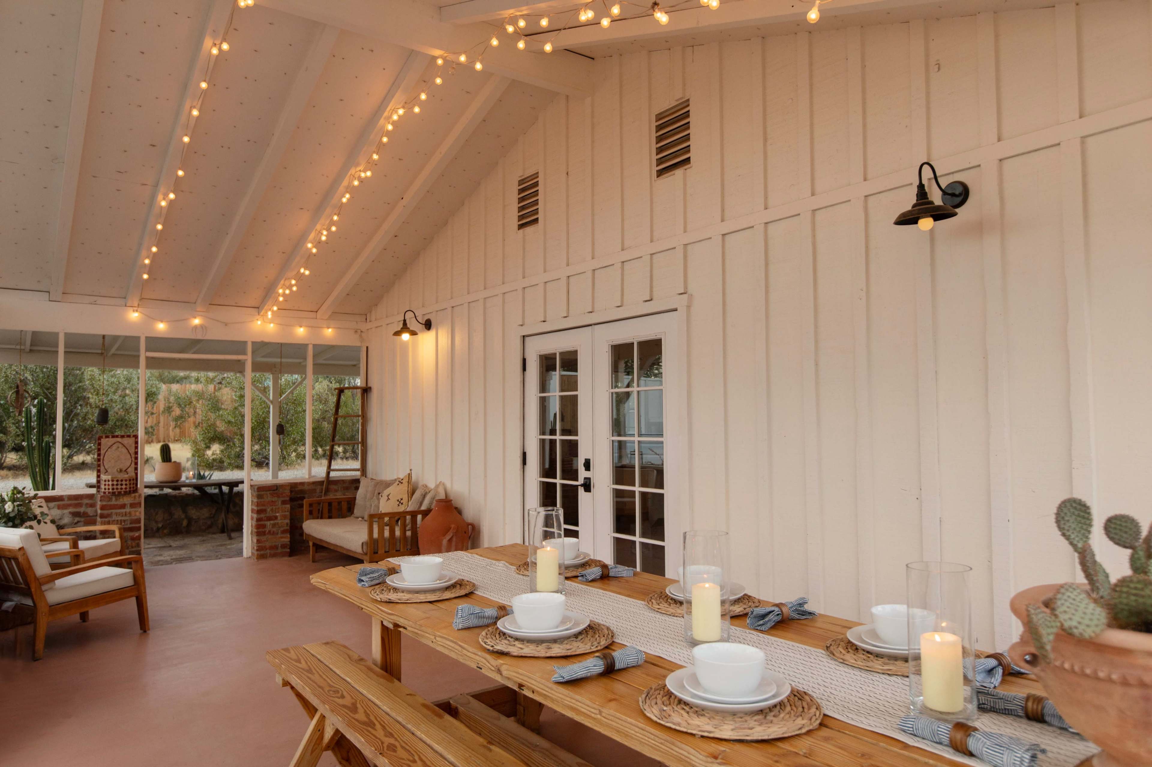 A well-lit outdoor dining area features a long wooden table set with plates and candles, surrounded by cozy seating and a view of greenery through large windows.