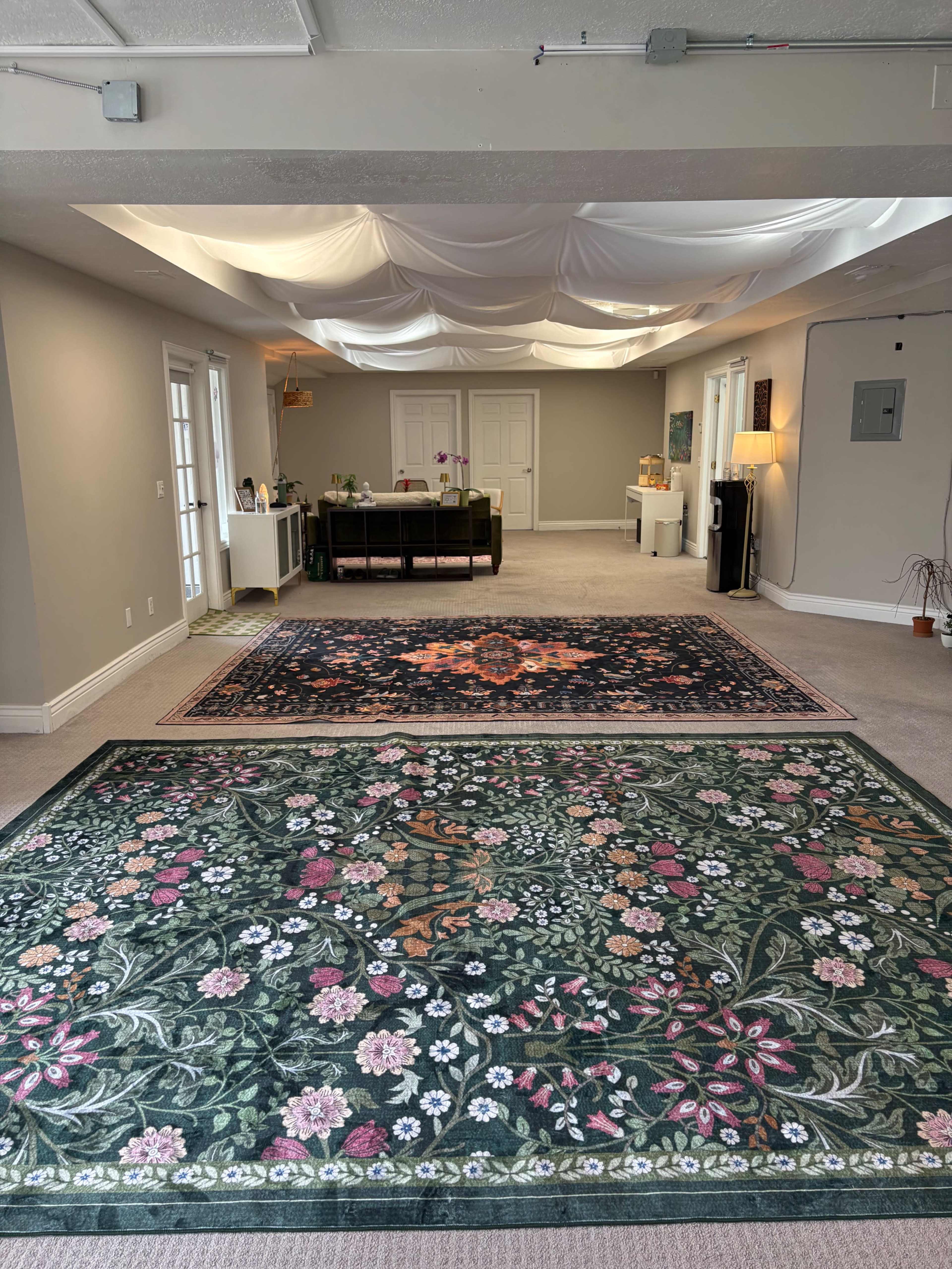 The image shows a spacious room featuring two large rugs on the floor, one with floral patterns and the other with a geometric design, and decorated with soft drapes on the ceiling.