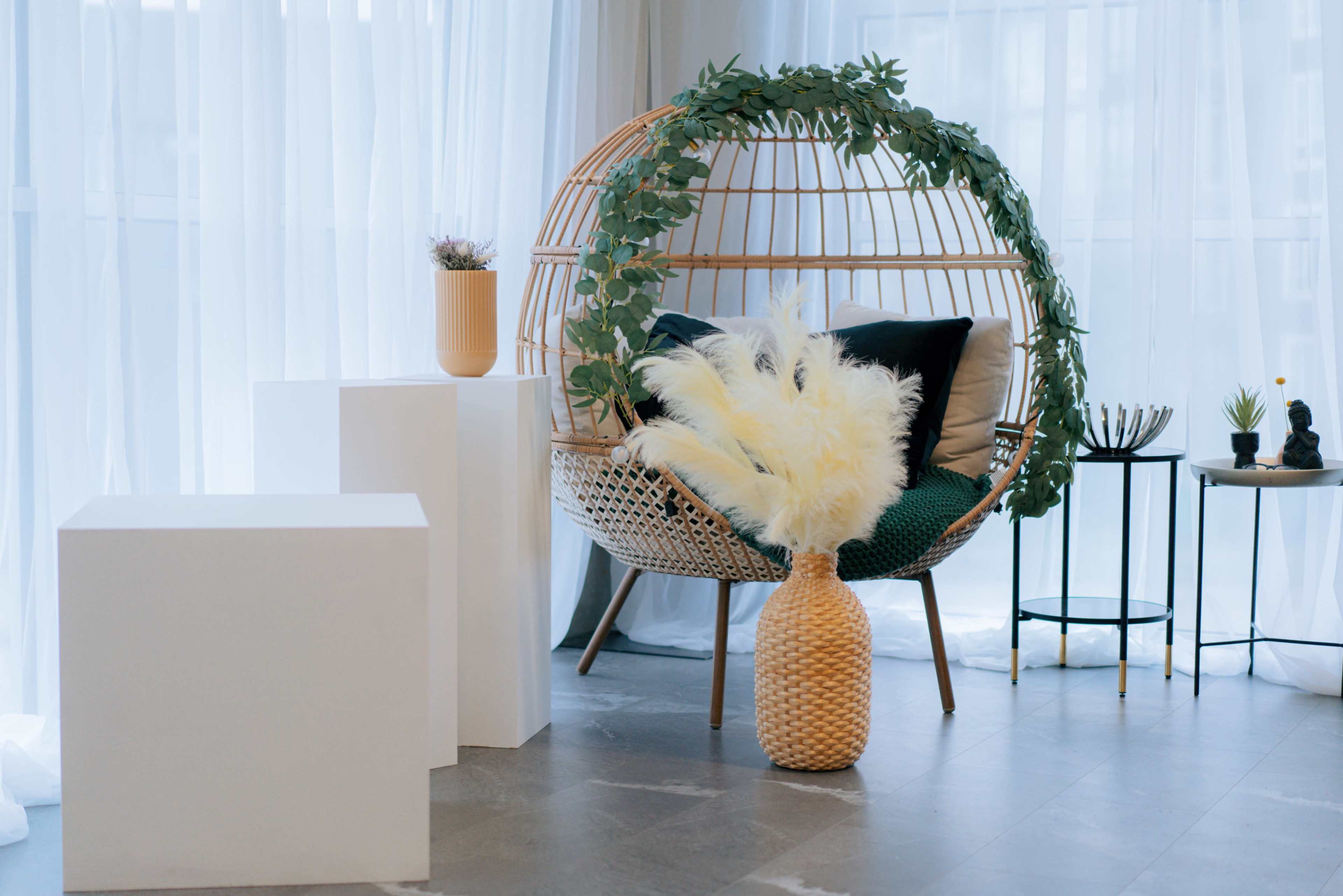 A stylish indoor seating area features a round rattan chair adorned with greenery, accompanied by modern pedestals and decorative vases on a tiled floor.