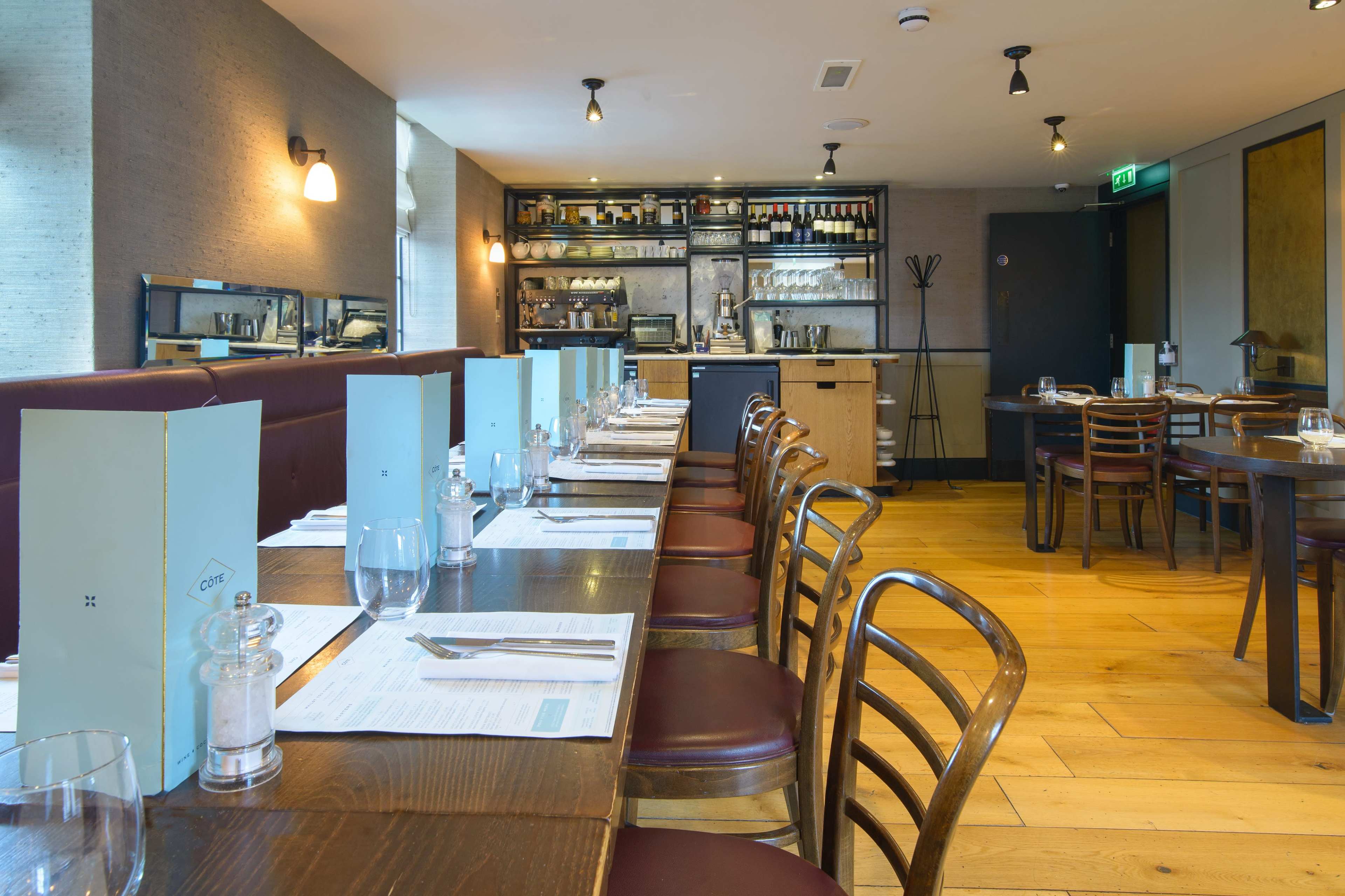 The image shows a modern restaurant interior with a long wooden table set for dining and a bar area in the background.