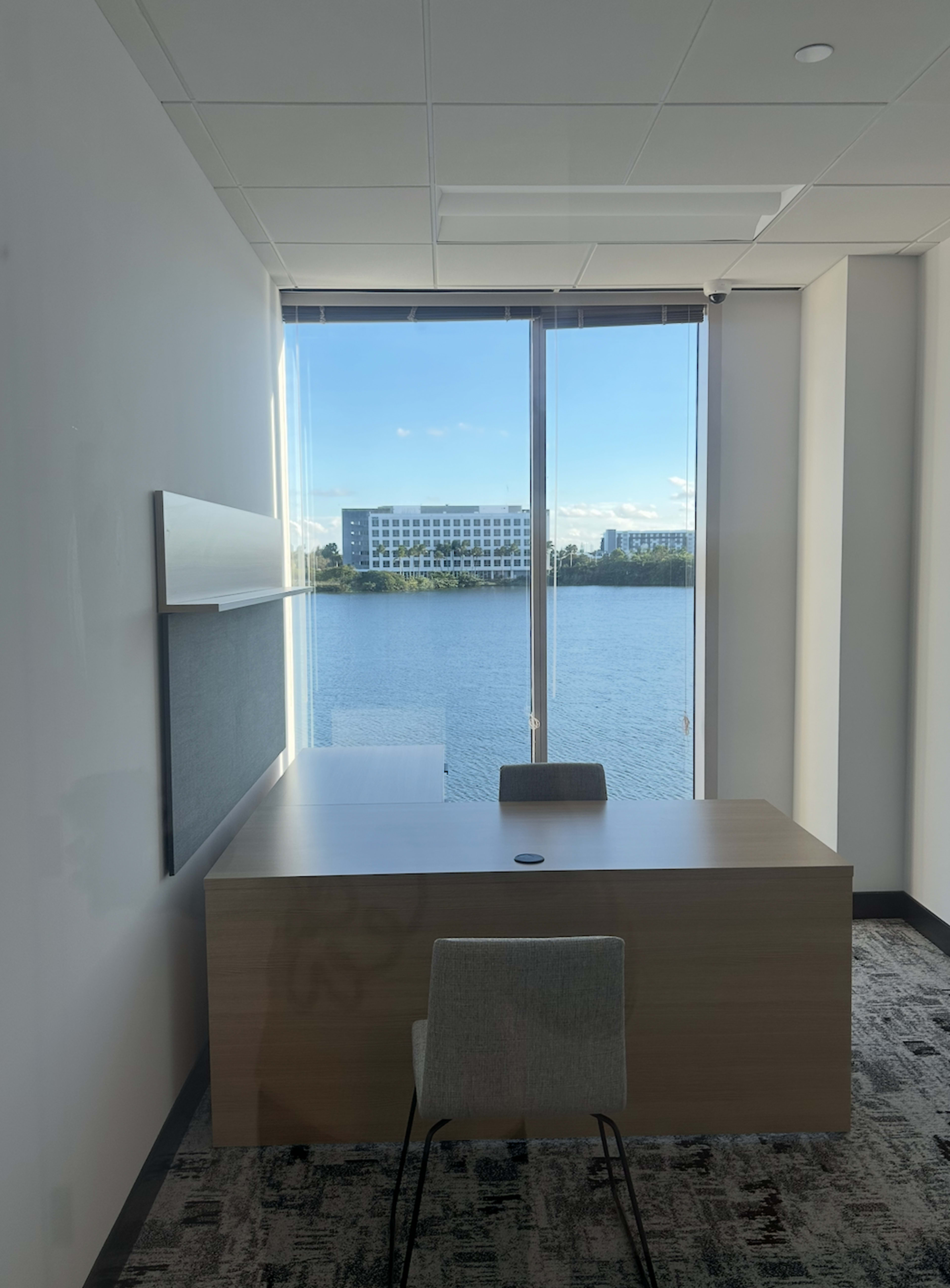 The image shows a modern office with a wooden desk and a chair, overlooking a body of water through large windows.