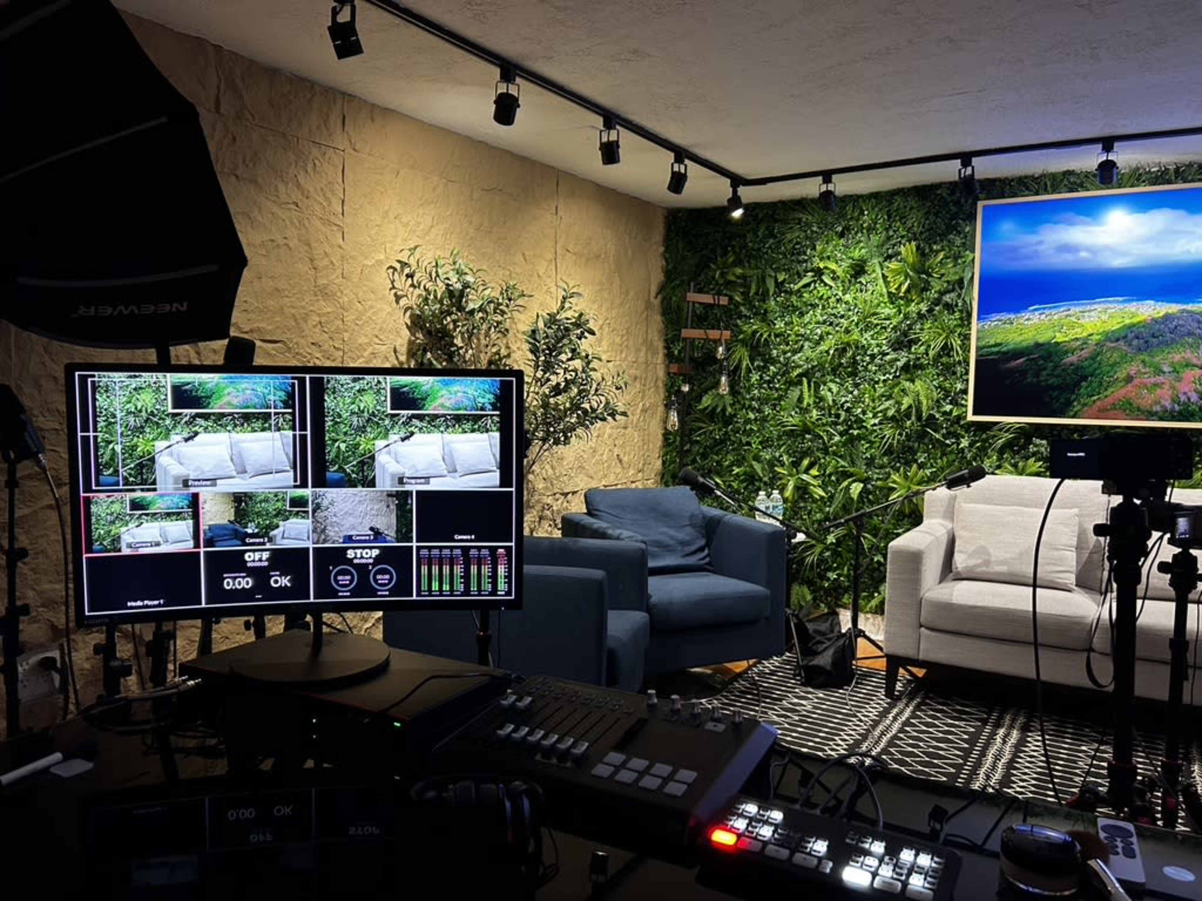 A television studio setup with multiple screens displaying video feeds, surrounded by greenery and two sofas against a textured wall.