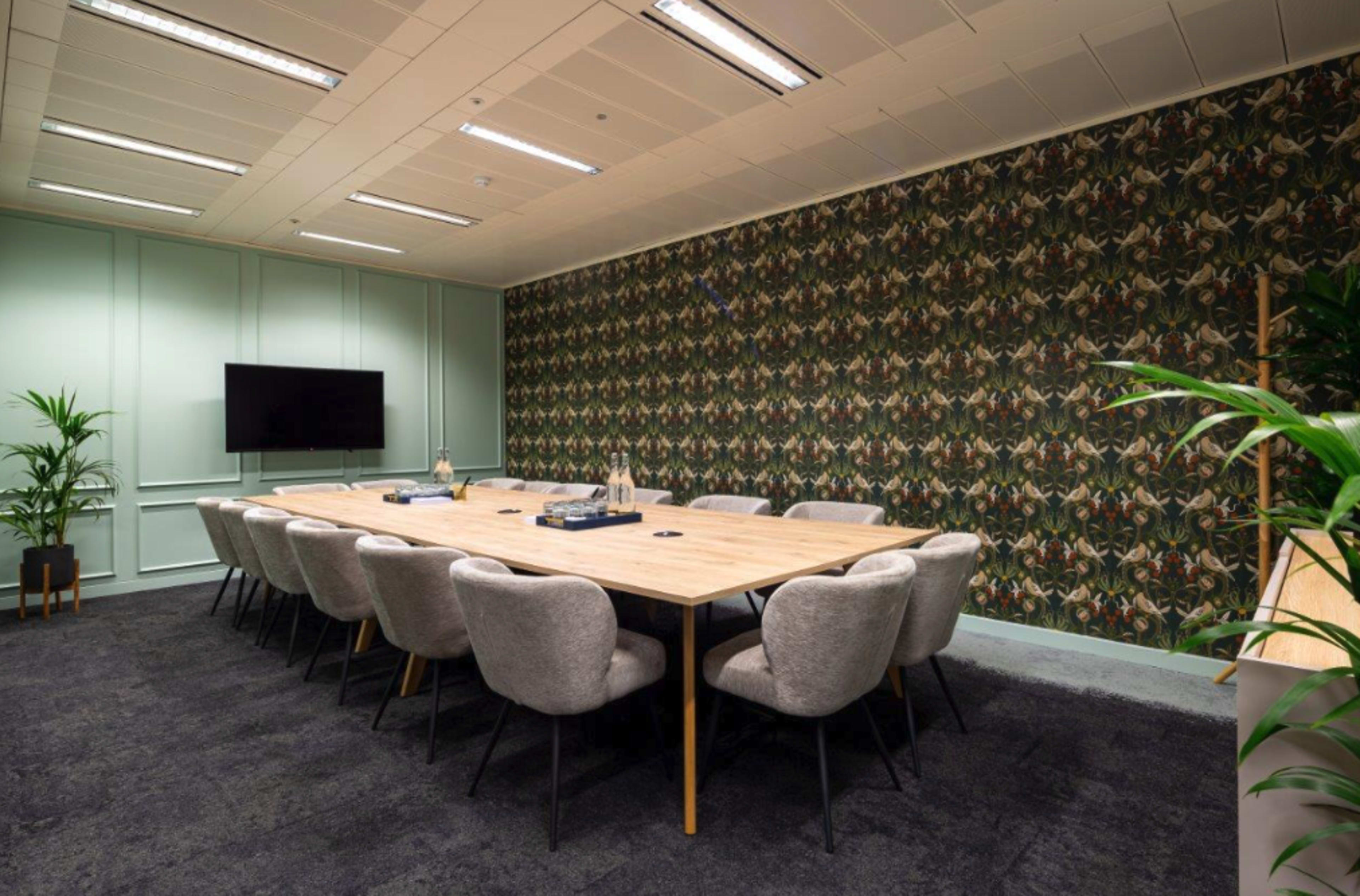 A modern conference room features a long wooden table surrounded by upholstered chairs, with decorative wallpaper and a television on one wall.