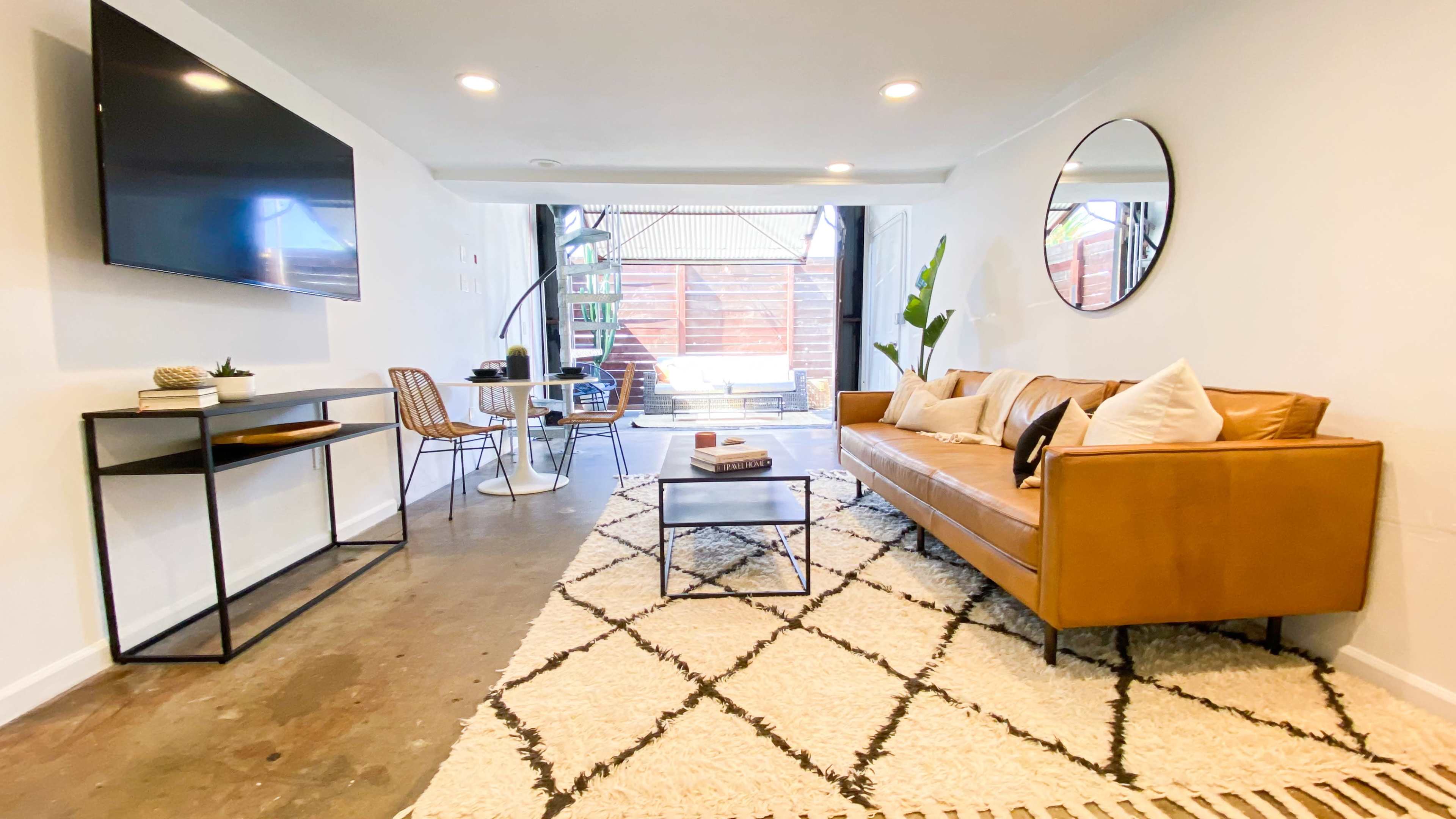 A modern living room with a brown leather sofa, a coffee table, a large wall-mounted TV, a round mirror, and a dining area featuring a small table and chairs.