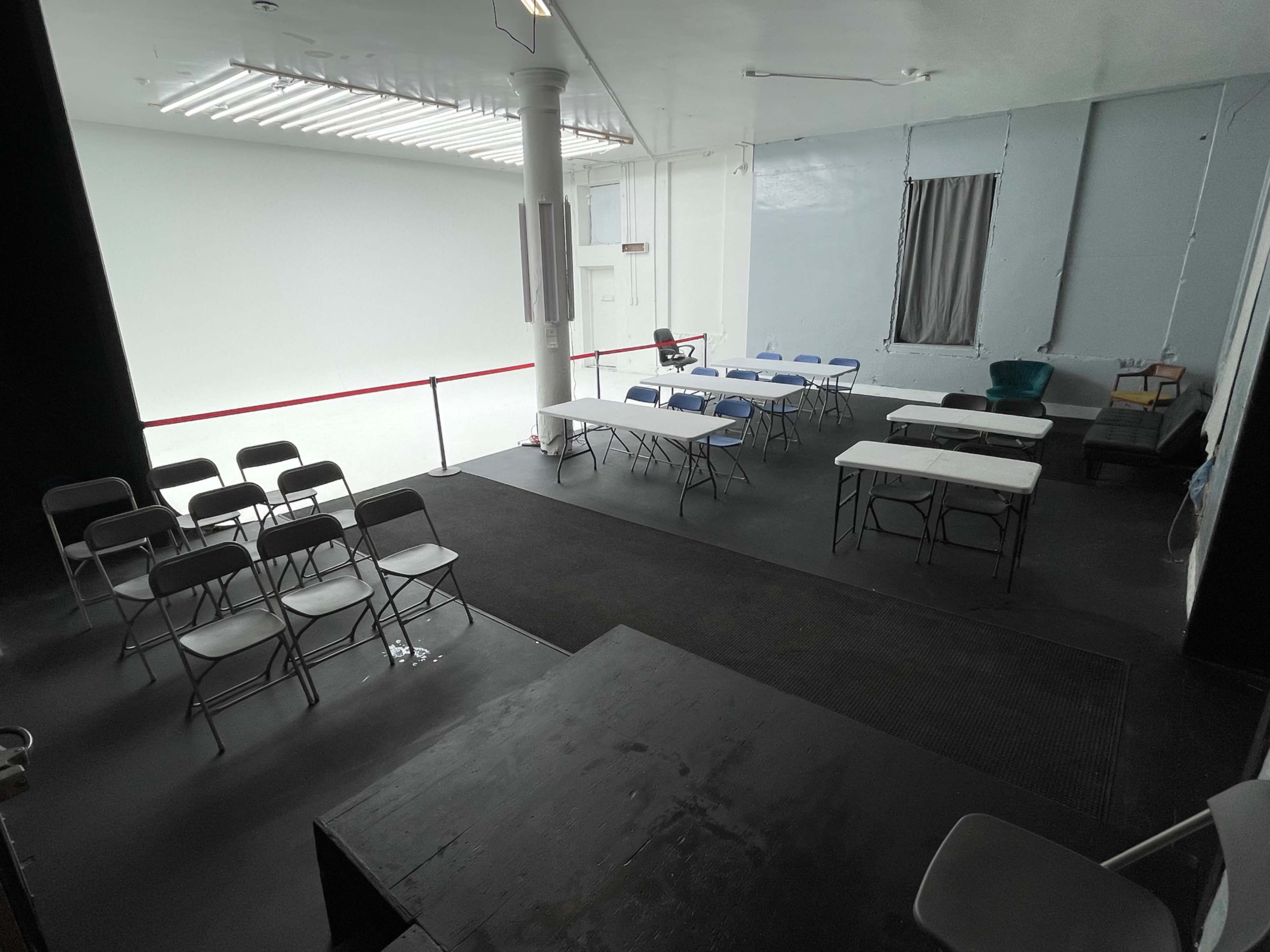 The image shows an empty room with multiple tables and chairs arranged in a setup for a meeting or event, with a white wall and a gray window in the background.