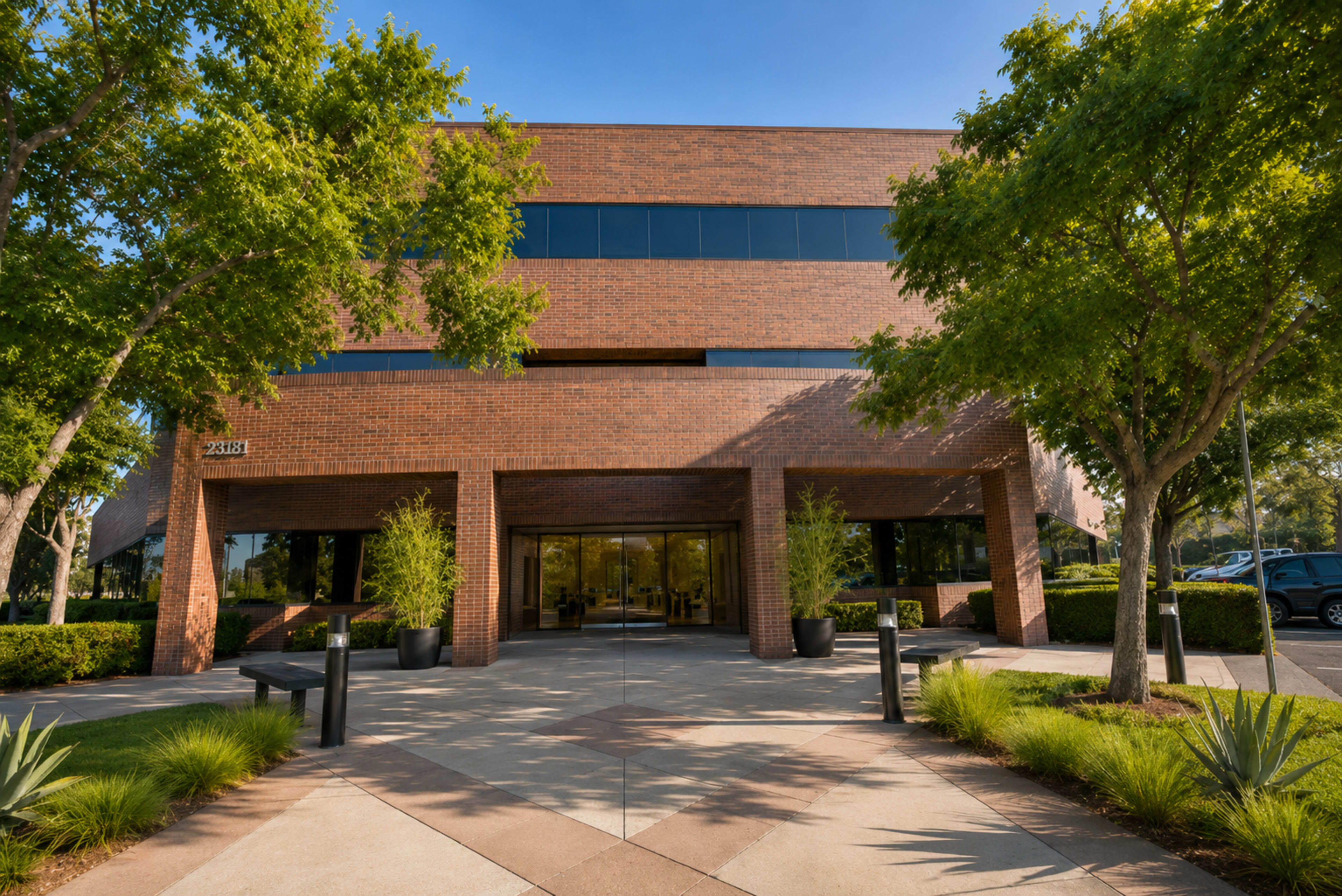 The image shows a modern brick office building with glass entrance surrounded by trees and landscaped greenery.