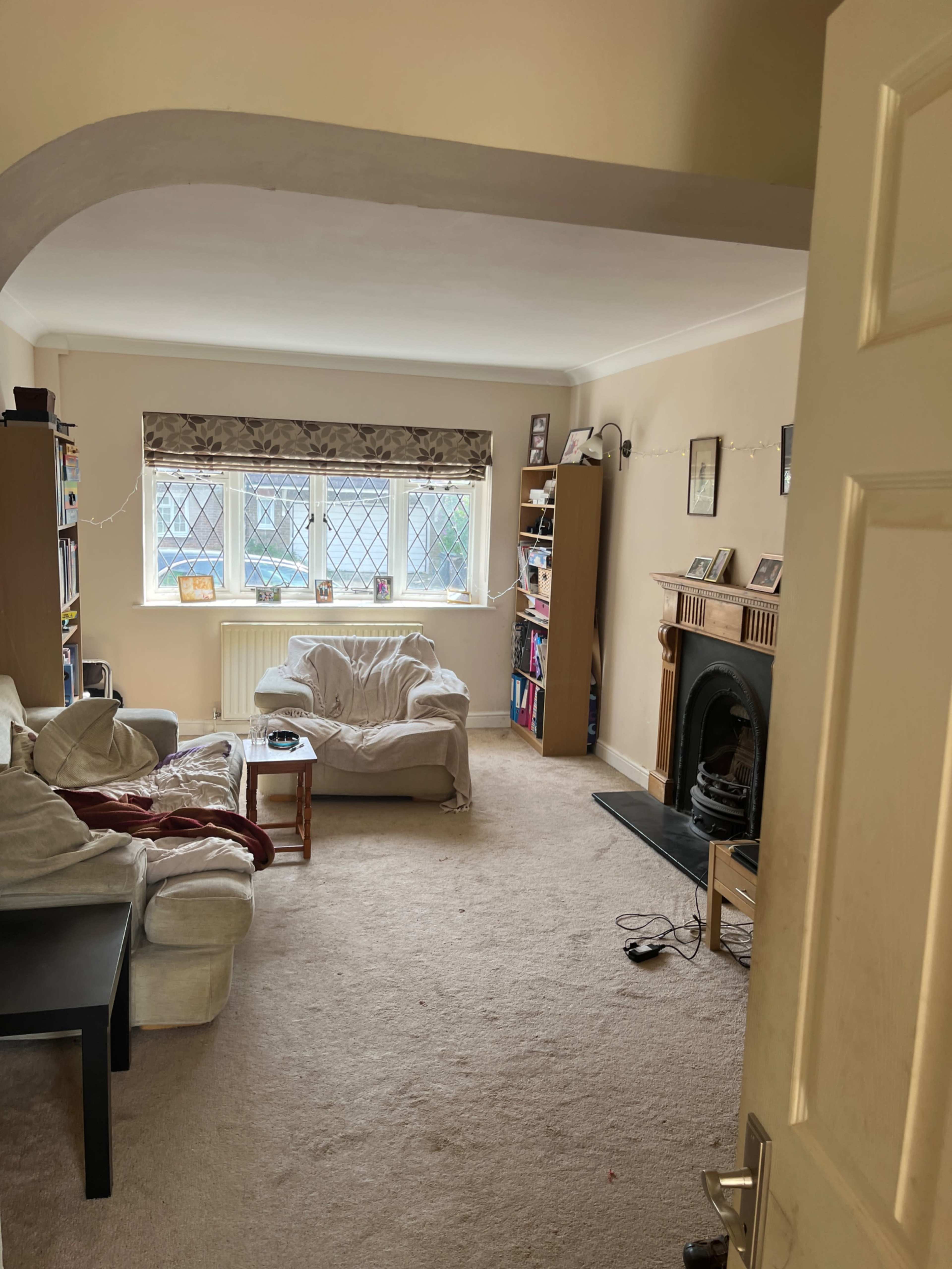 The image shows a living room with a light-colored carpet, two sofas, a coffee table, bookshelves, and a fireplace, viewed from an open door.