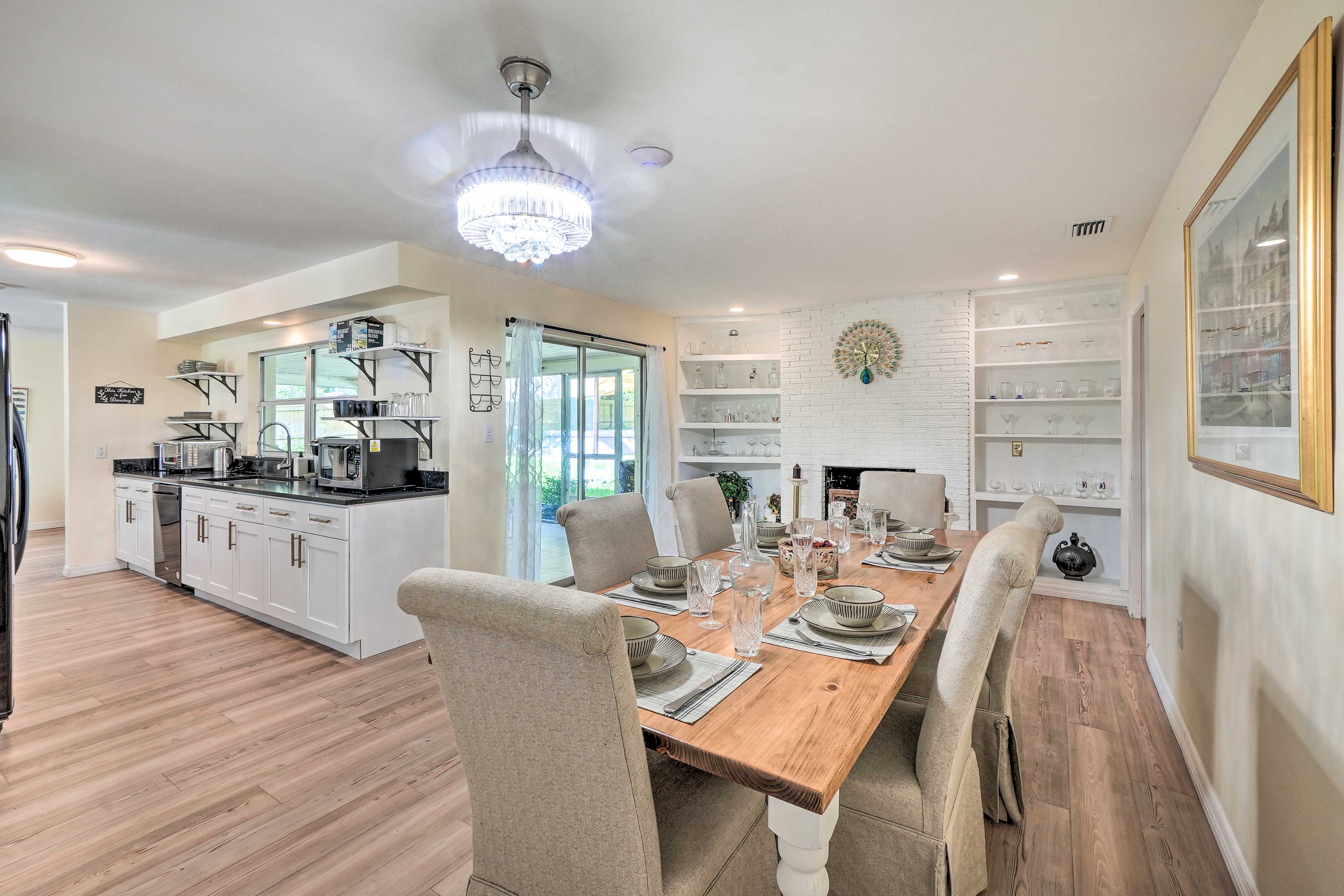 A dining room setup with a wooden table laid for a meal, surrounded by upholstered chairs, and a modern kitchen area with white cabinetry and open shelving in the background.