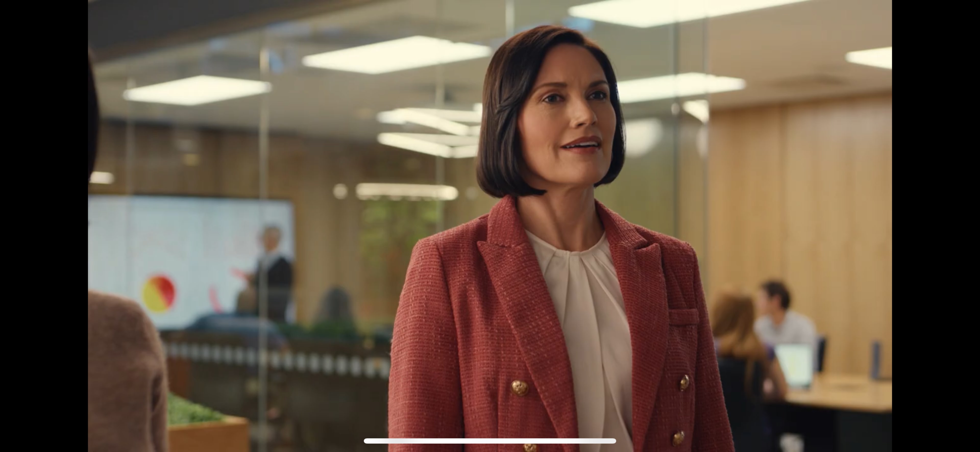 A woman in a tailored blazer stands in an office with glass walls, while a presentation is being given in the background.