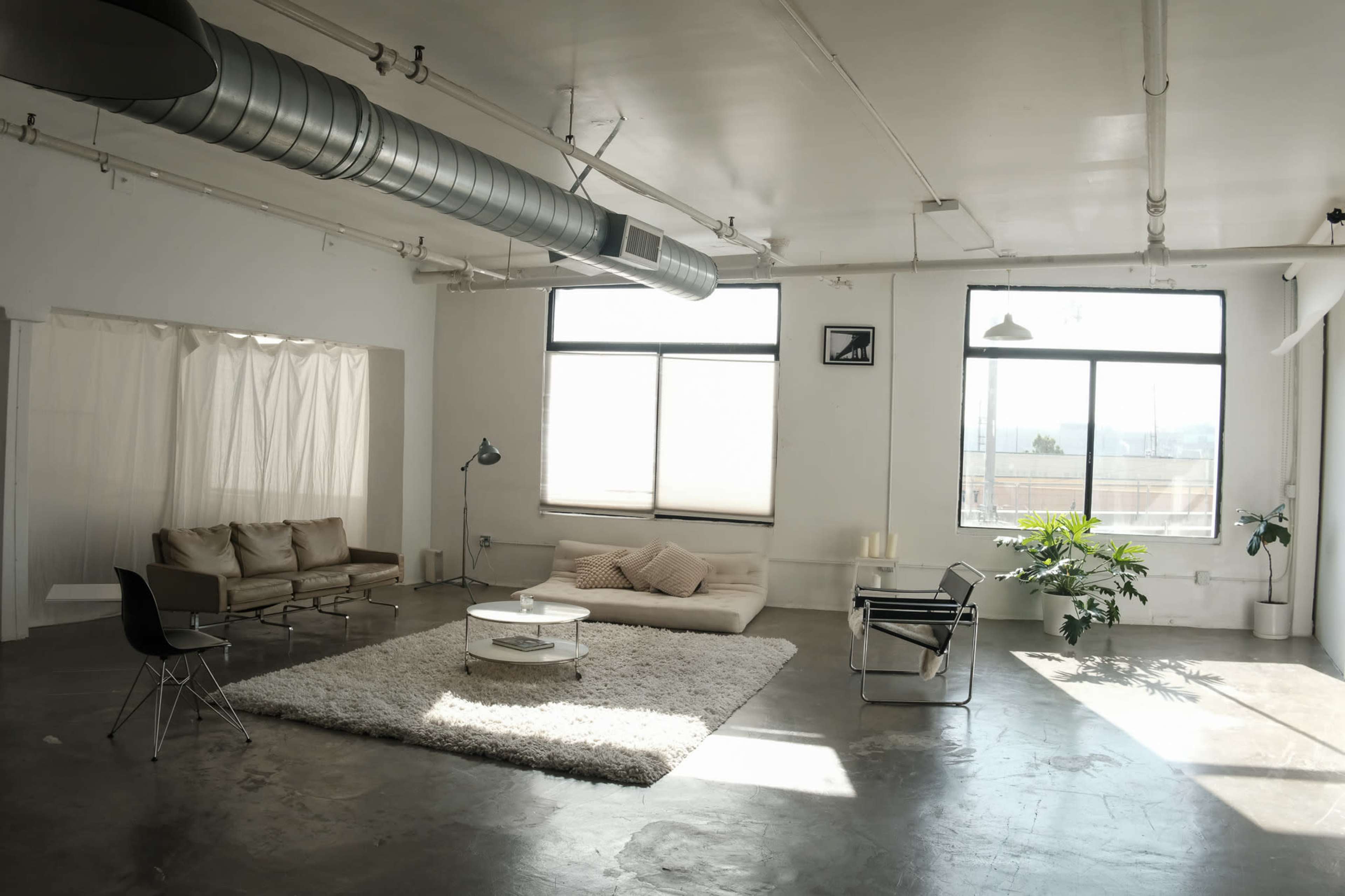 The image shows a spacious, minimally furnished industrial-style living room with large windows, a beige sofa, a coffee table, and a rug on a concrete floor.