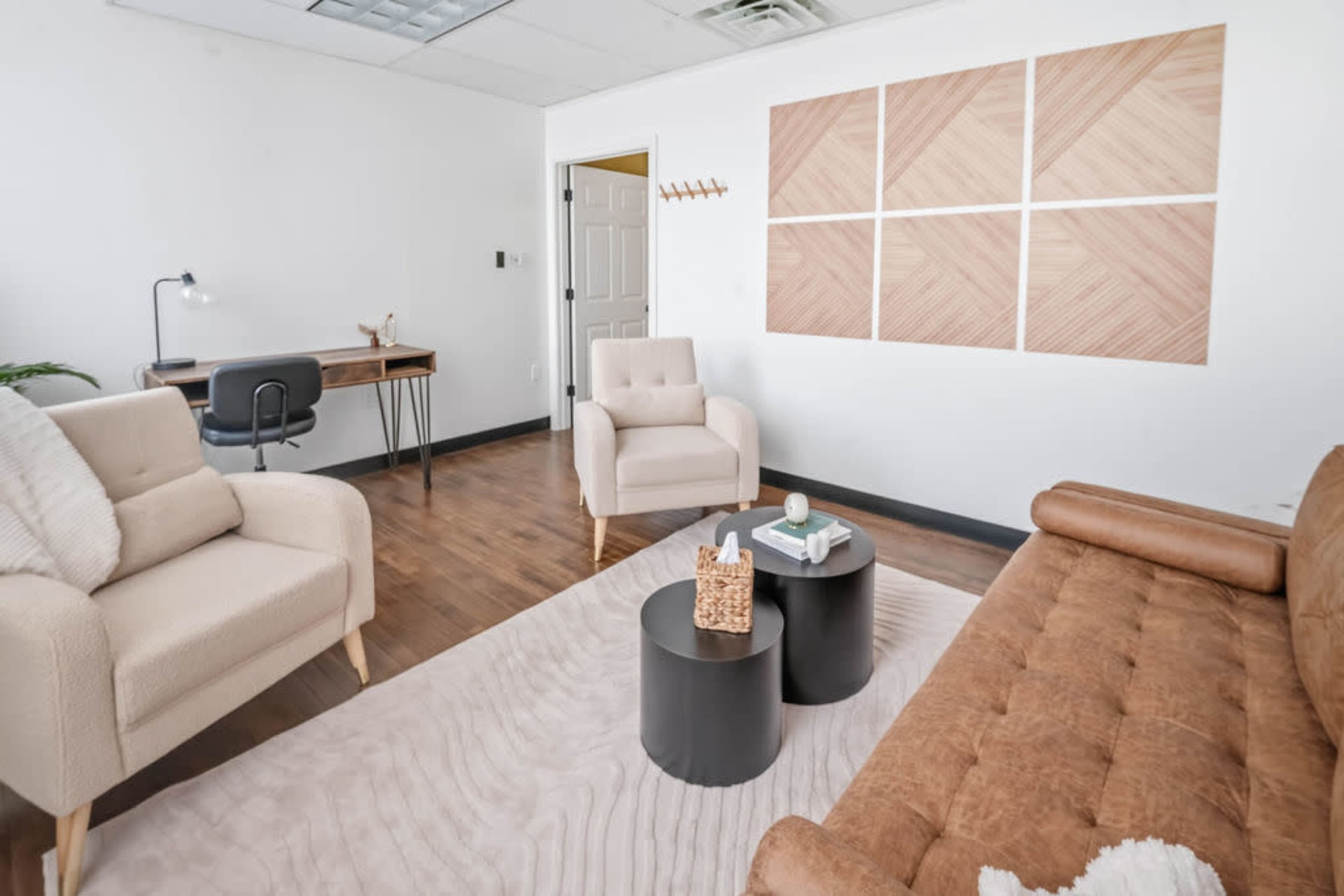 The image shows a minimalist office space featuring two comfortable chairs, a brown sofa, a small black coffee table, and a wooden desk against a white wall with geometric artwork.