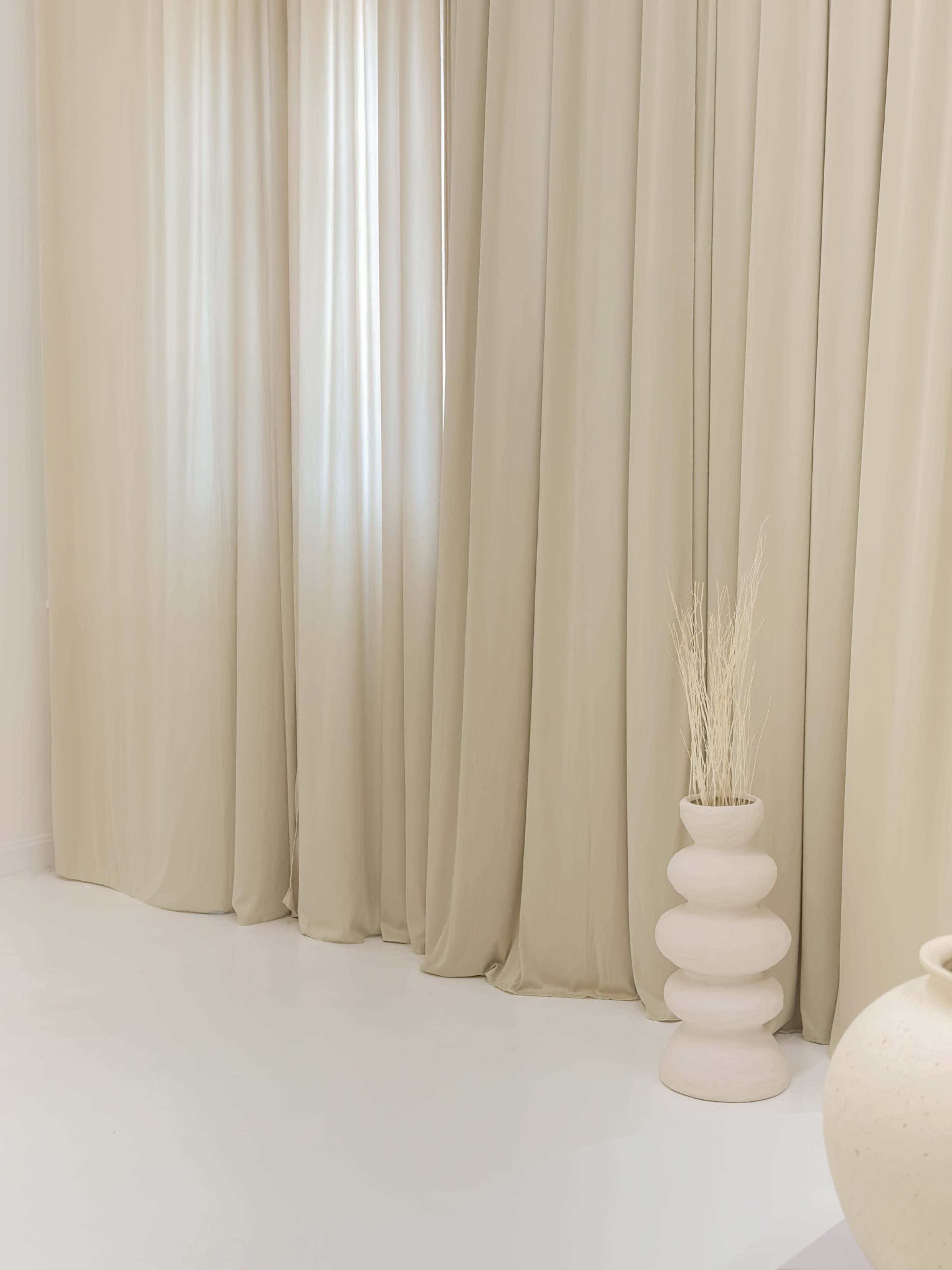 A light-colored room with long, draping curtains and a decorative ceramic vase containing dried plants.