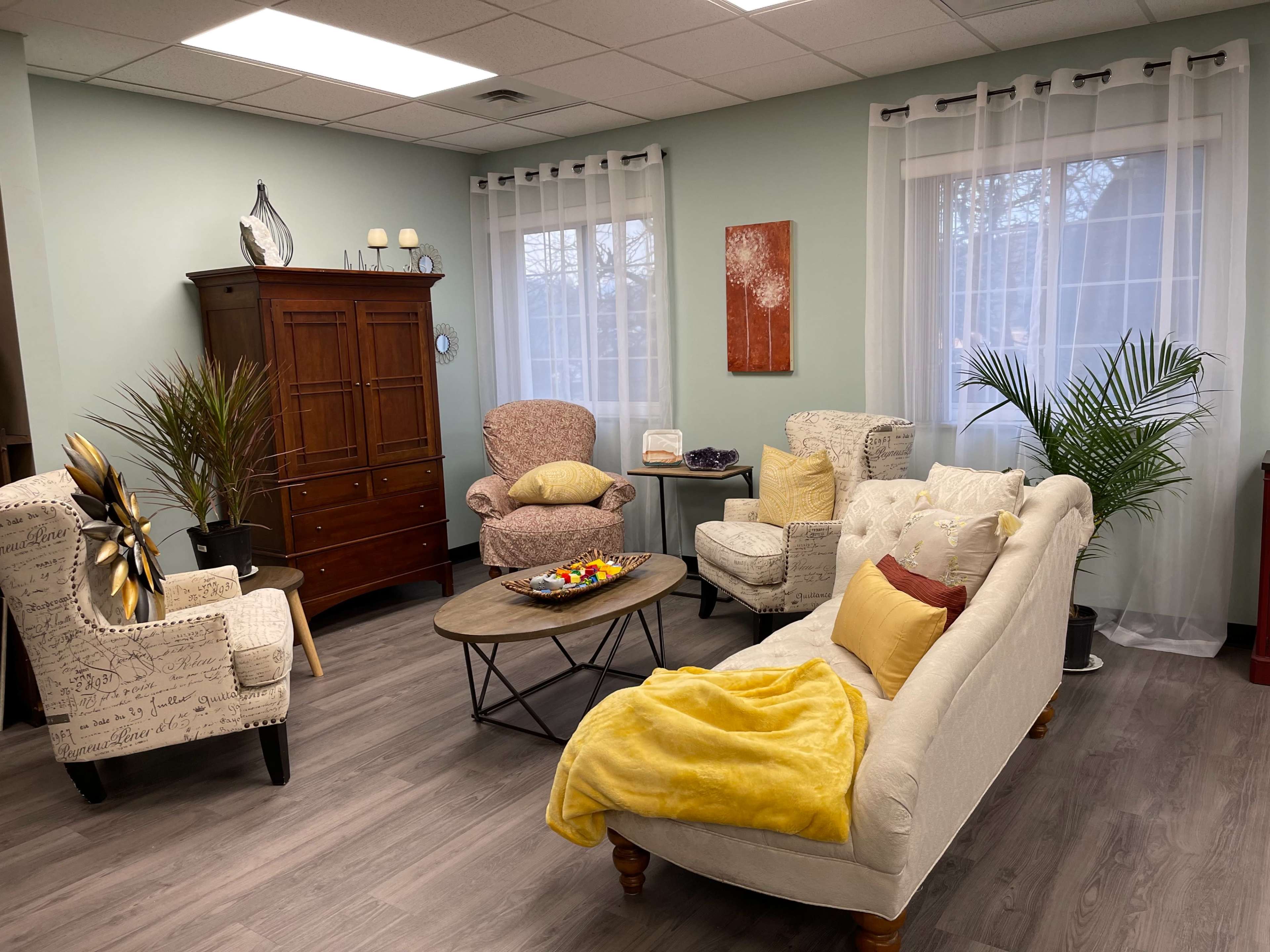 The scene depicts a cozy living room with two upholstered chairs, a beige couch, and a wooden coffee table, surrounded by potted plants and natural light from large windows.