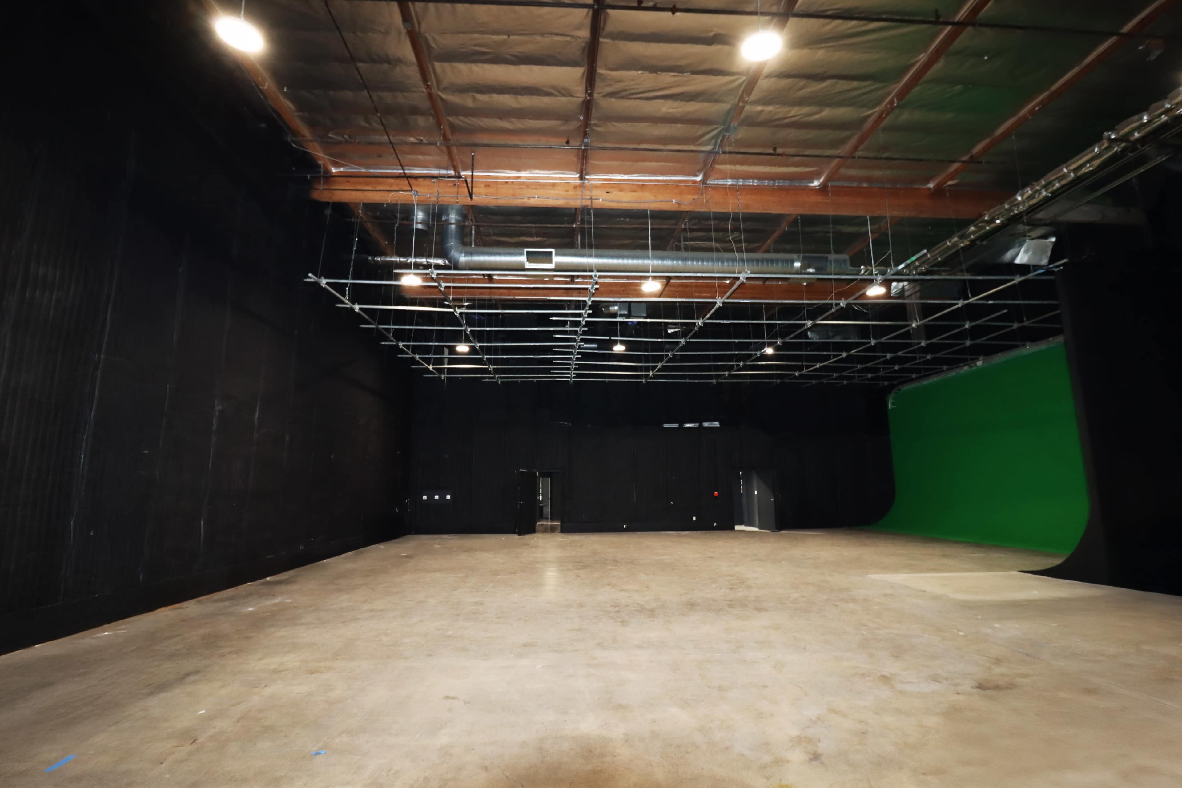 The image shows an empty studio space with black walls, a concrete floor, and a green screen at one end.