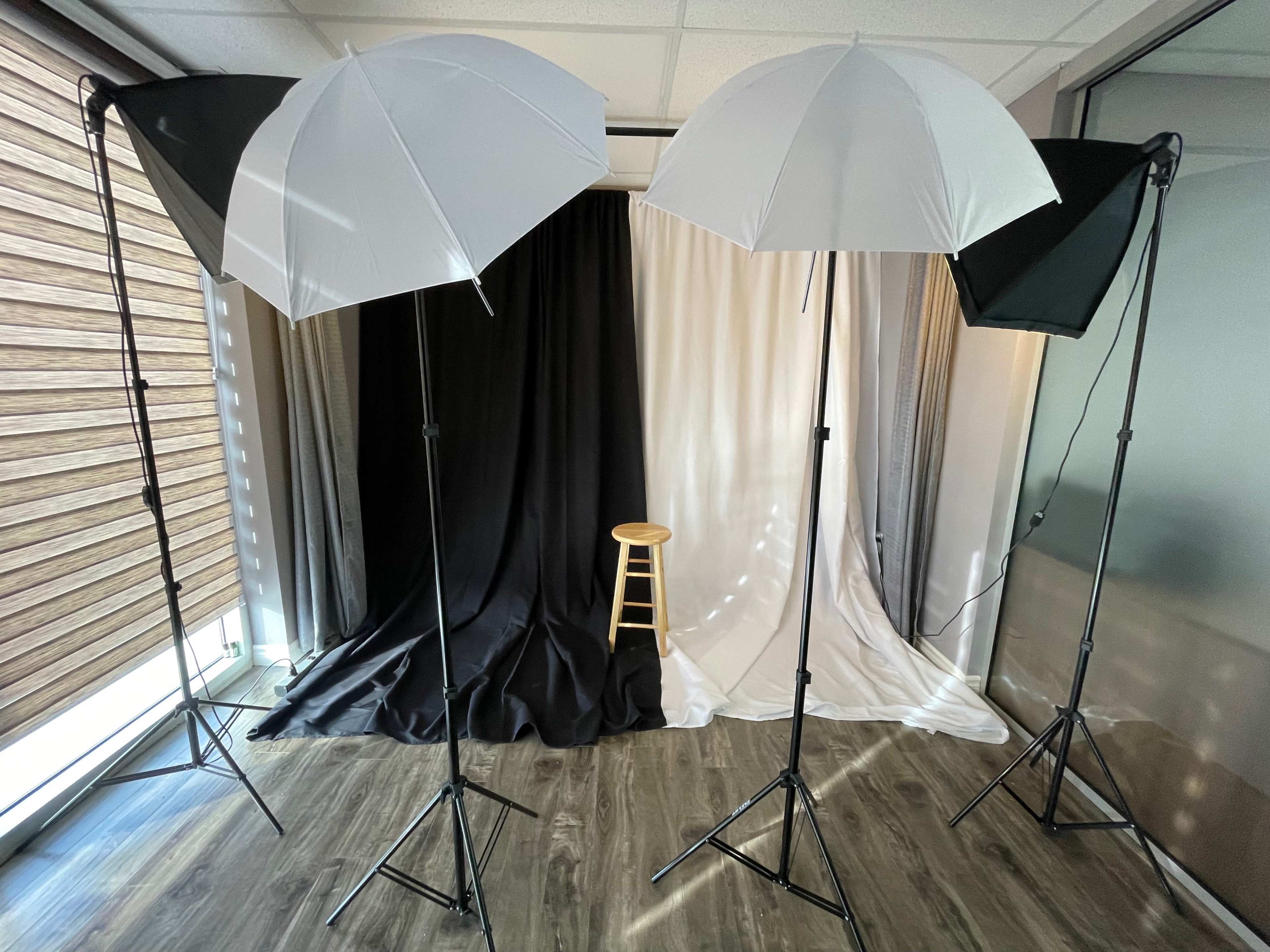A well-lit photography setup with two umbrella lights on stands facing a three-panel backdrop of black, white, and gray, with a wooden stool positioned in front.